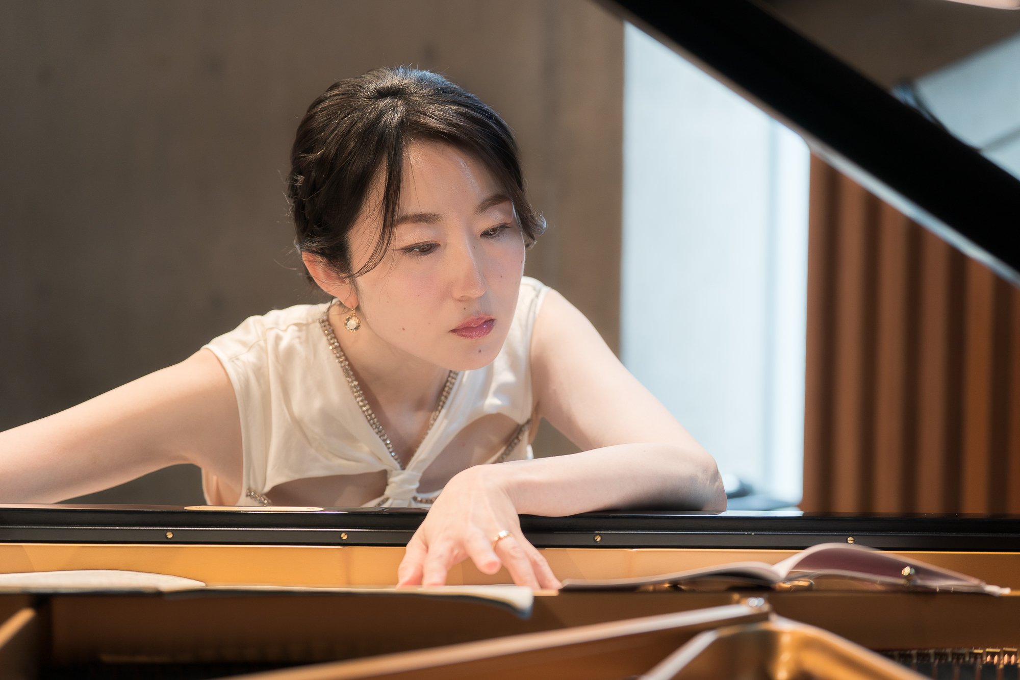 A woman with dark hair styled in a short bob wearing a sleeveless white top and gold earrings is playing the piano, focusing on the keys as she looks down carefully.