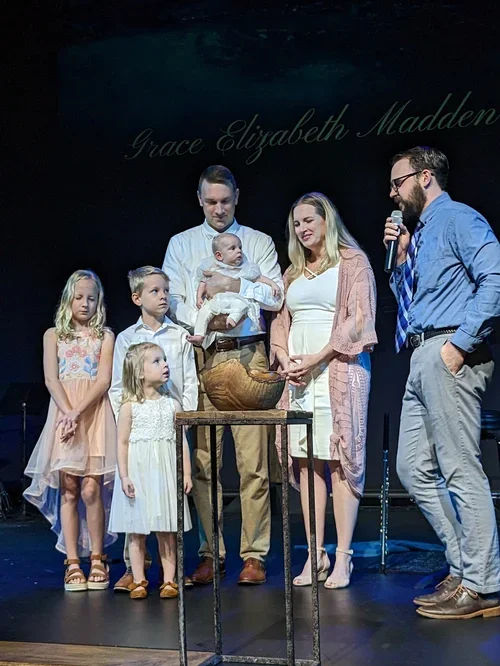 A family participating in a christening or baptism ceremony on stage, with a pastor or officiant, and children present. The backdrop has the name Grace Elizabeth Madden.