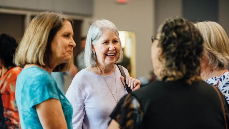Group of women conversing at an indoor social event, smiling and engaging with each other.