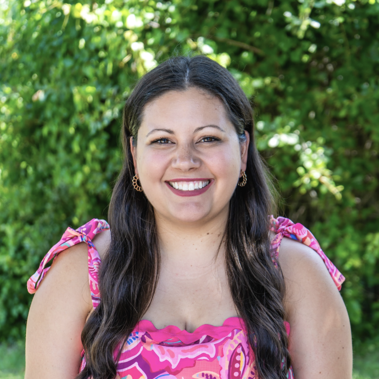 Smiling woman outdoors with green foliage in background, wearing pink dress with ruffled neckline and bow details on shoulders, gold hoop earrings.
