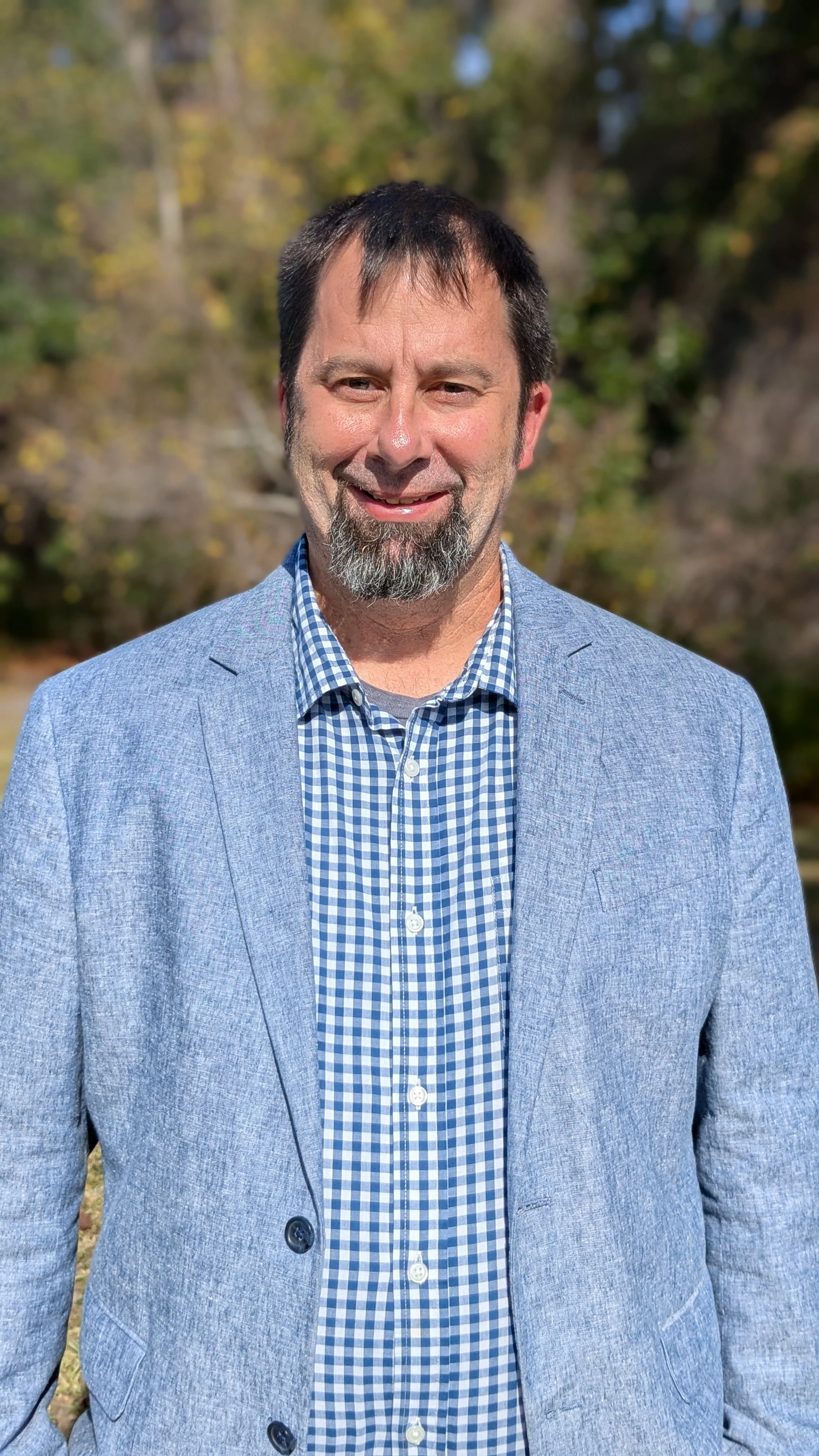 A man smiling outdoors, wearing a blue checked shirt and a light blue blazer, with a blurred background of trees and greenery.