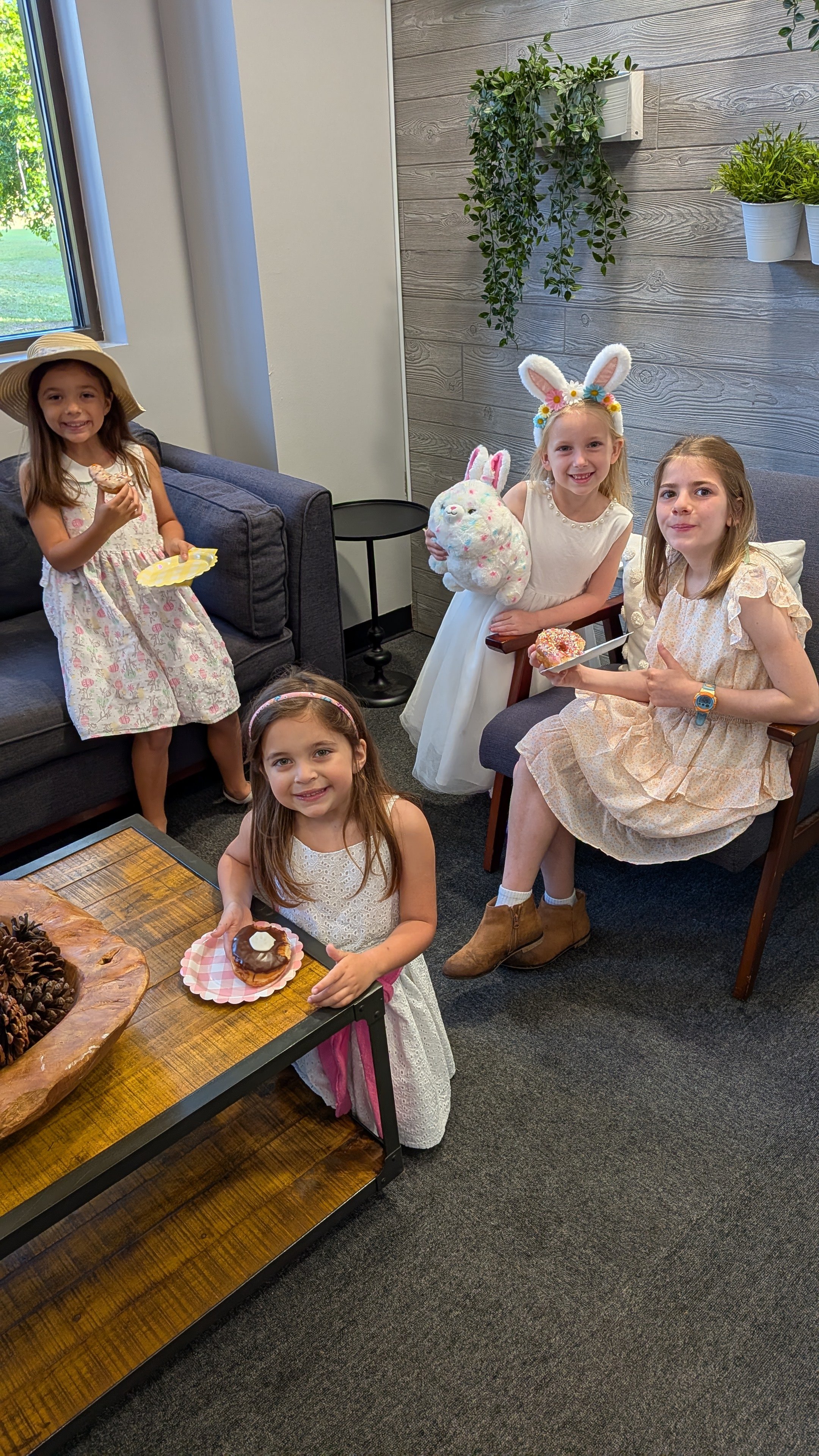 Five young girls at a birthday celebration, some wearing Easter bunny headbands, holding plates with desserts in a room with a wooden wall and potted plants.