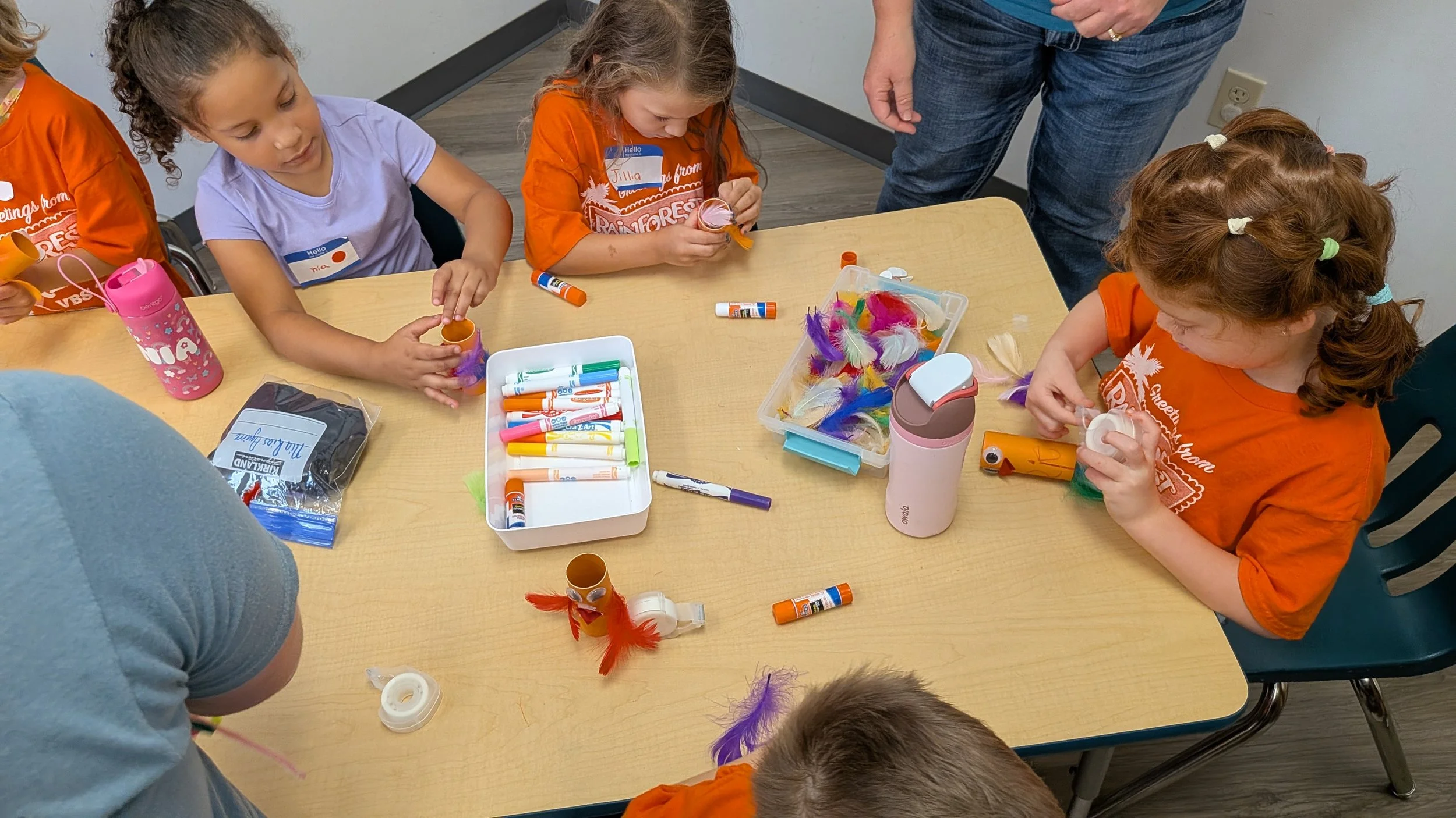 Several young children with curly and straight hair engaged in an arts and crafts activity at a table. They are using glue sticks, ornaments, and colorful feathers for decorating. An adult is overseeing the activity.
