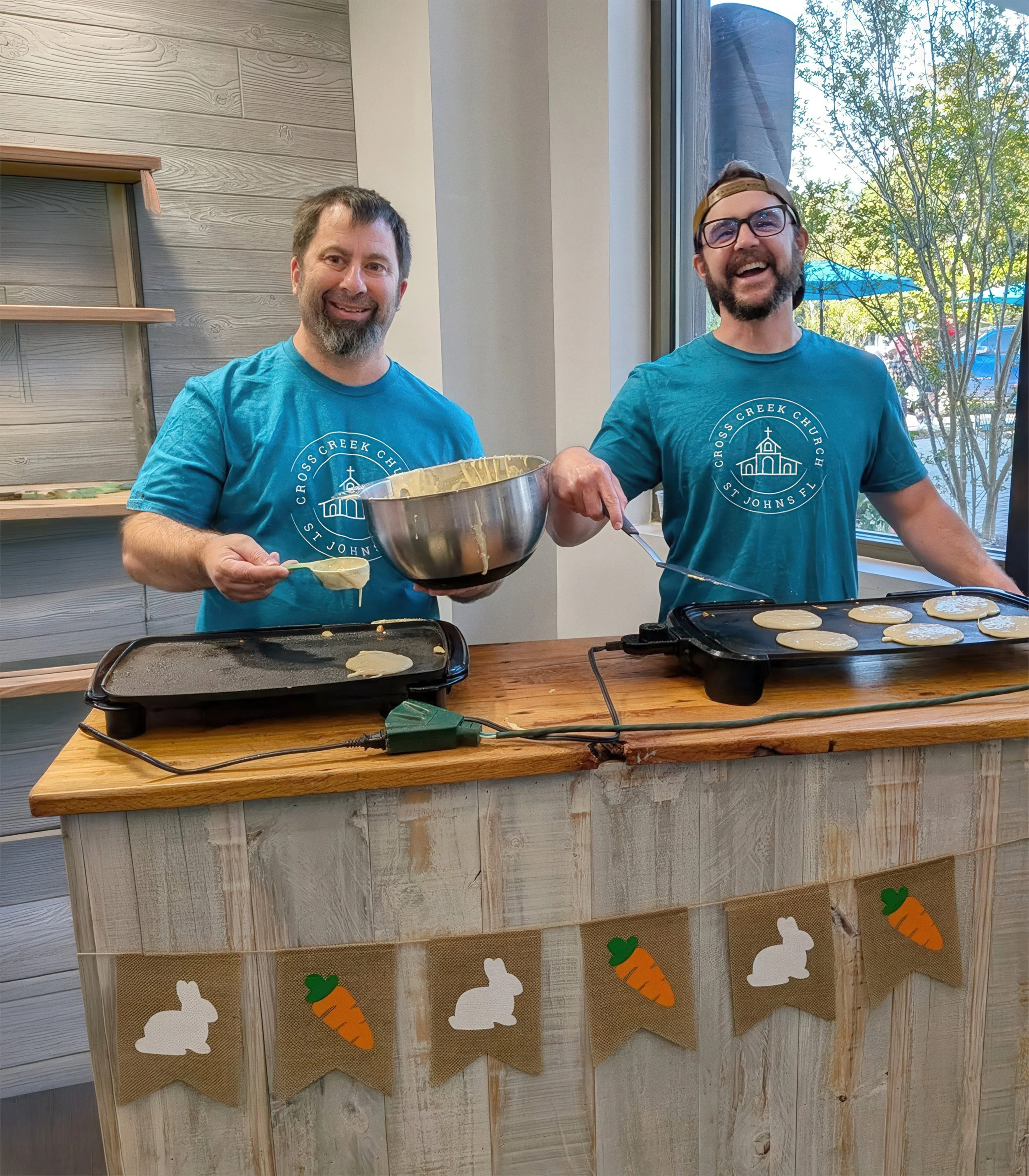 Two smiling men in blue Cross Creek Church t-shirts, one holding a bowl of batter, standing behind a table with electric griddles cooking pancakes. Decorated with paper carrots and bunnies on a banner underneath a window, inside a room with wooden walls.