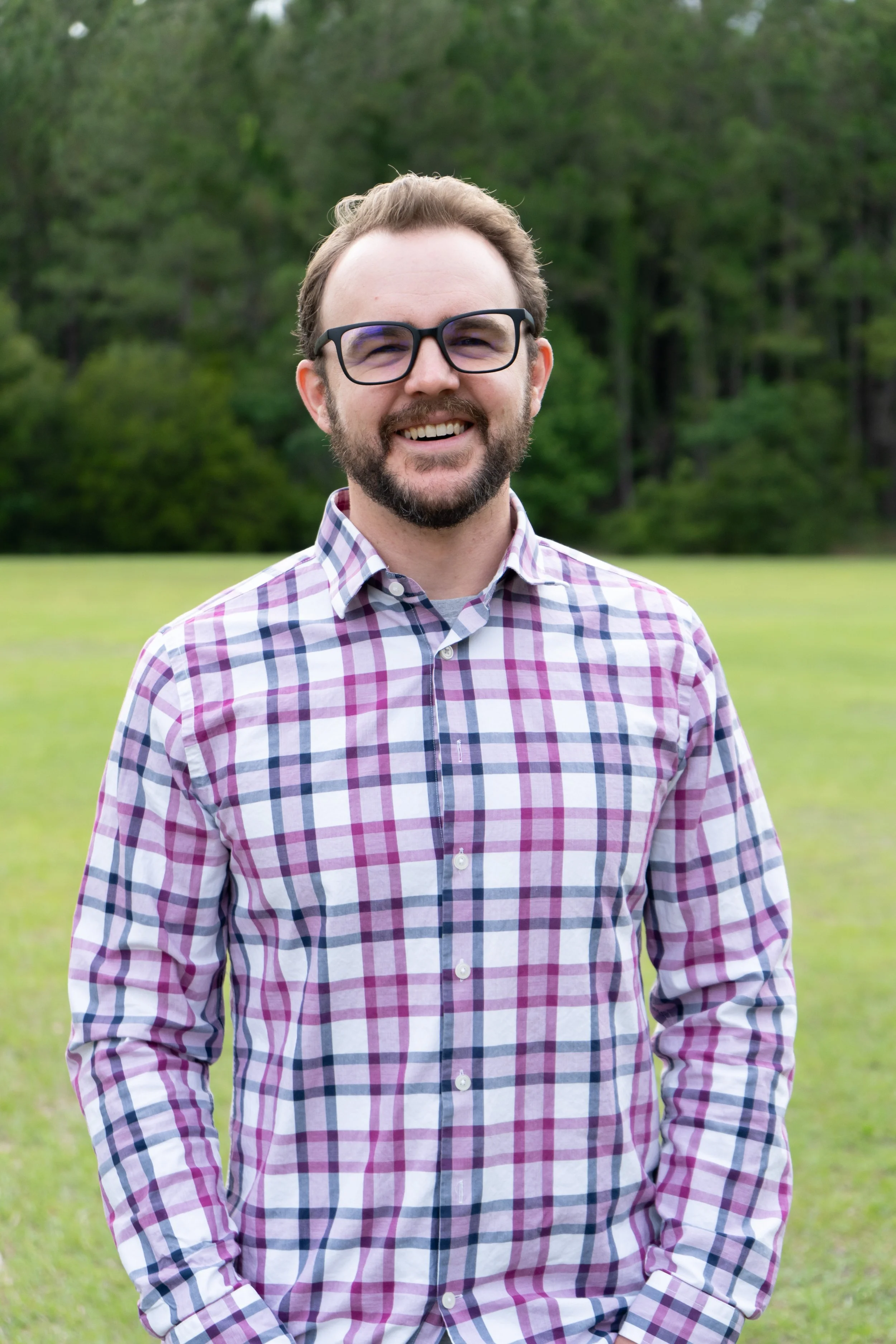 A man with glasses and a beard smiling outdoors in front of green trees and grass.