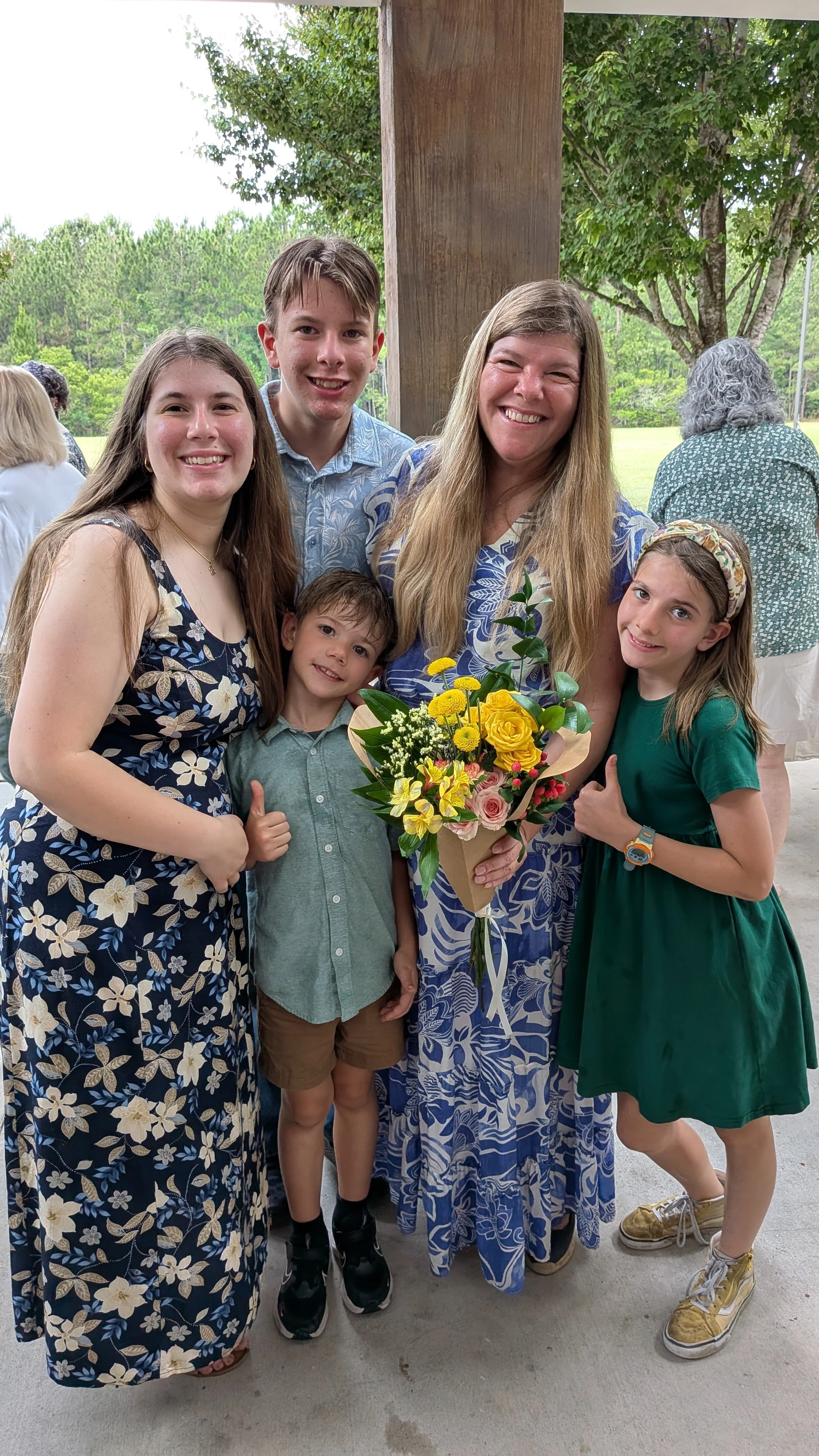 A group of six people, including three young children and three adults, standing outdoors near a wooden structure, smiling, with trees and grass in the background. One of the adults holds a bouquet of flowers.