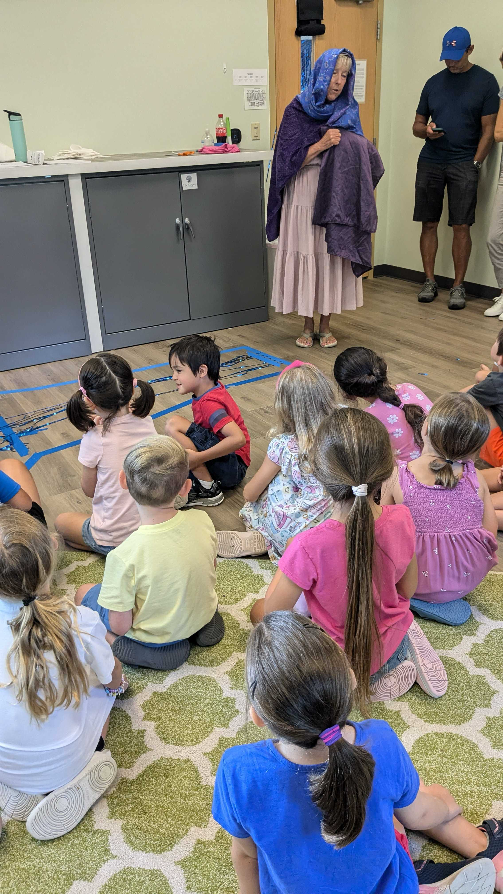 A woman dressed in a costume with a purple shawl and white dress leading a group of children in an indoor room. The children are sitting on a green patterned carpet, listening attentively.