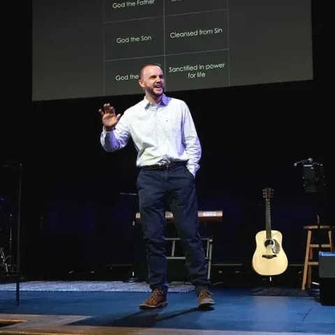 A man is standing on a stage, speaking or presenting with a slide behind him that lists religious references. He is smiling, wearing a white shirt, navy pants, and brown shoes. There is a guitar on a stand and a stool on the stage.