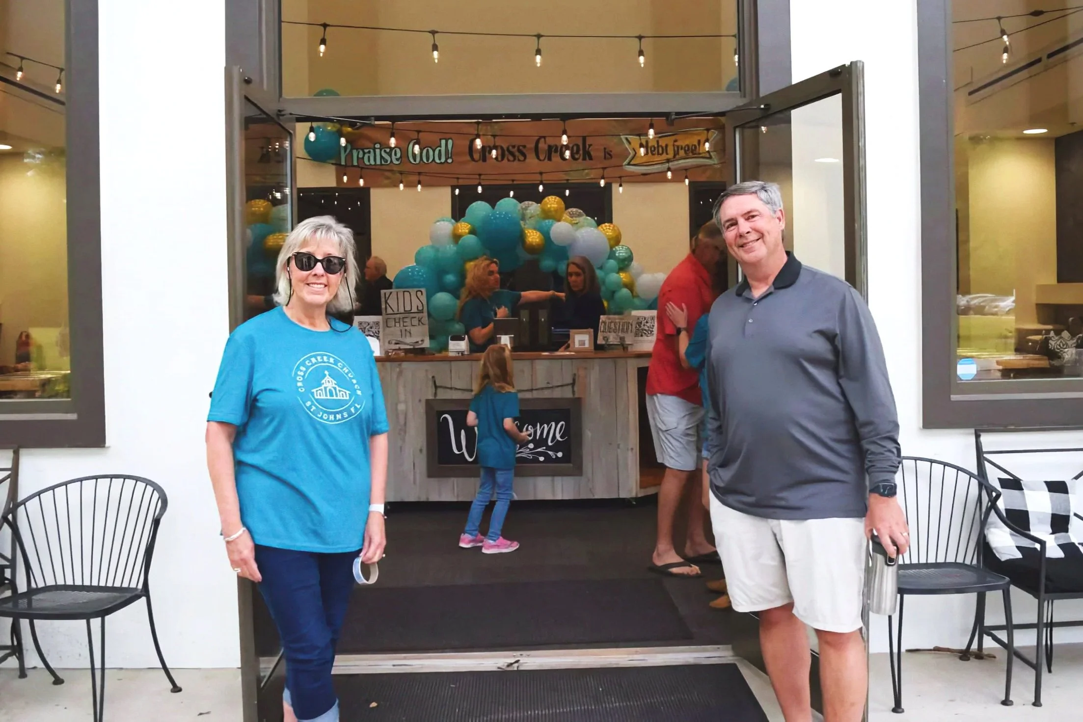 Two smiling adults, a woman wearing sunglasses and a blue T-shirt and a man in a gray button-up shirt, standing outside a church entrance with a decorated sign and balloons, welcoming visitors.