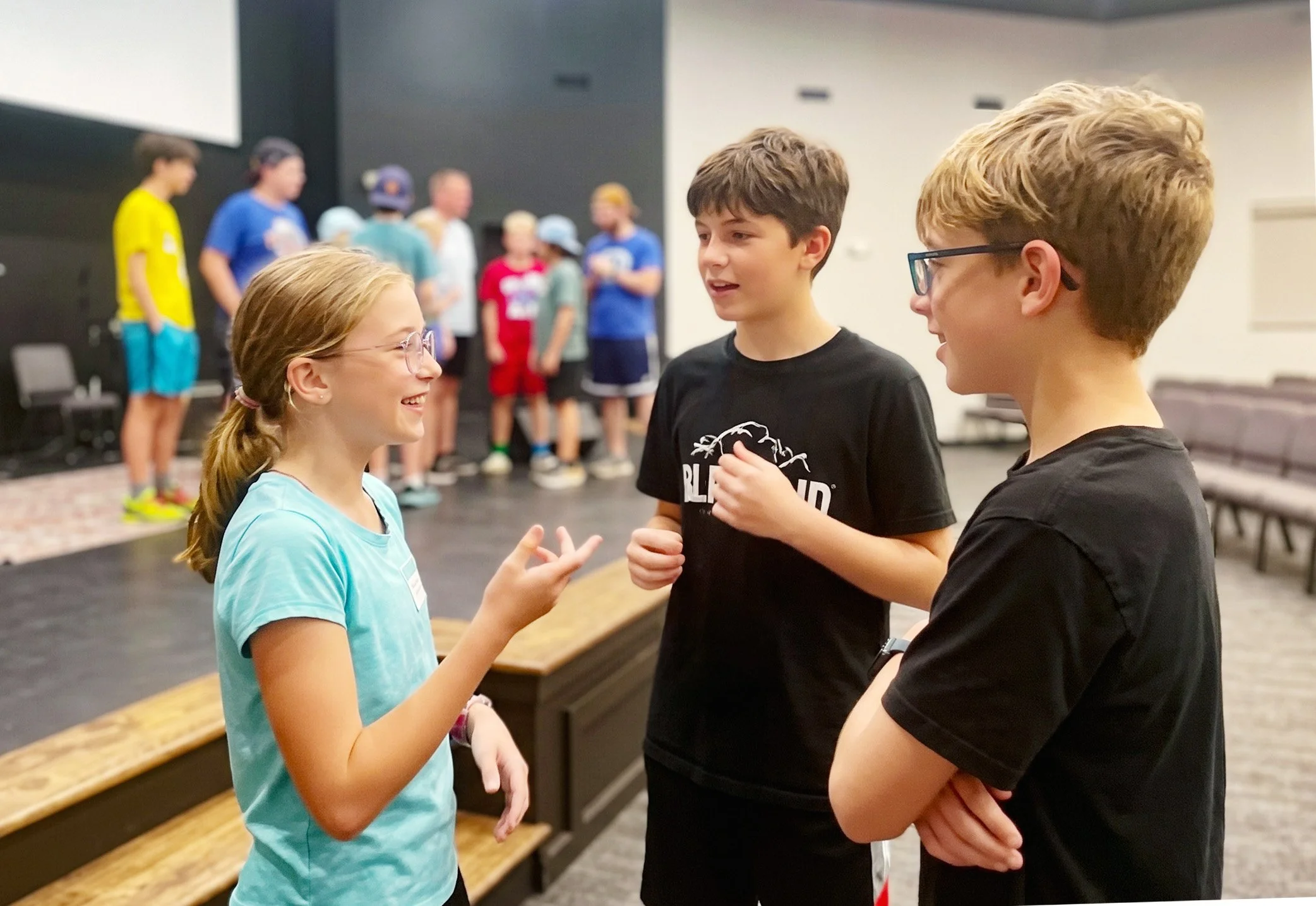 Three children are engaged in conversation in a room. Two boys and a girl, all smiling, are talking to each other. In the background, several other children are standing together, some wearing colorful t-shirts and hats, with blurred faces indicating a group setting.