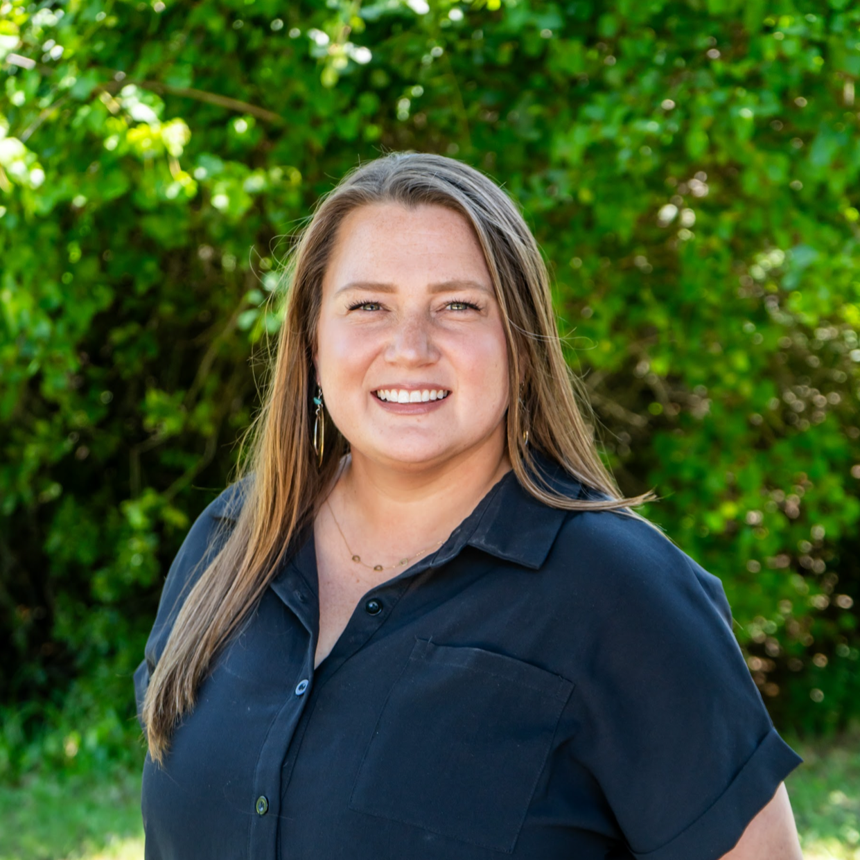 A woman with long, light brown hair smiling outdoors in front of green foliage, wearing a black button-up shirt and gold jewelry.