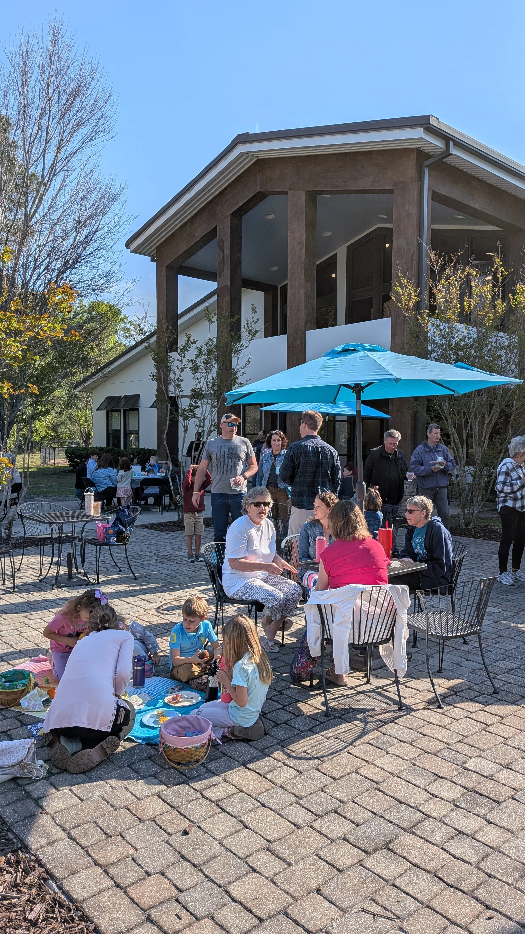People gathered outdoors for an event, sitting and standing around tables with umbrellas, near a modern building with large windows and wooden accents, on a sunny day.