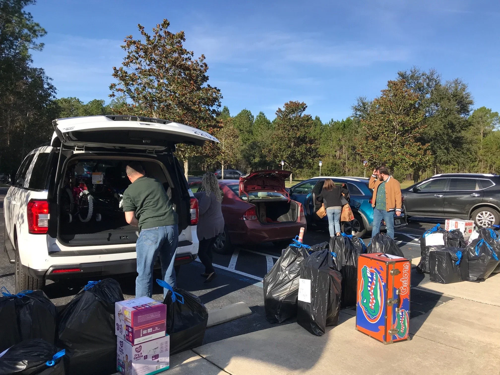 People organizing and packing donations in a parking lot under a clear blue sky, with several parked cars and trees in the background.