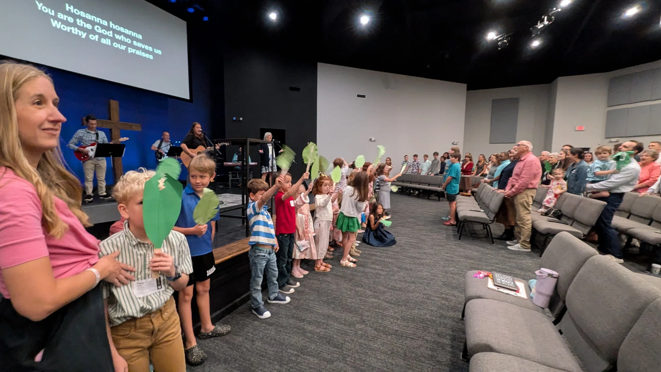 People, including children and adults, participating in a church service or gathering inside a church auditorium. Children are standing in front, holding green paper leaves. Musicians are playing instruments on stage in the background. A large screen displays lyrics to a song titled "Hosanna Hosanna." The congregation is engaged, some standing and singing.