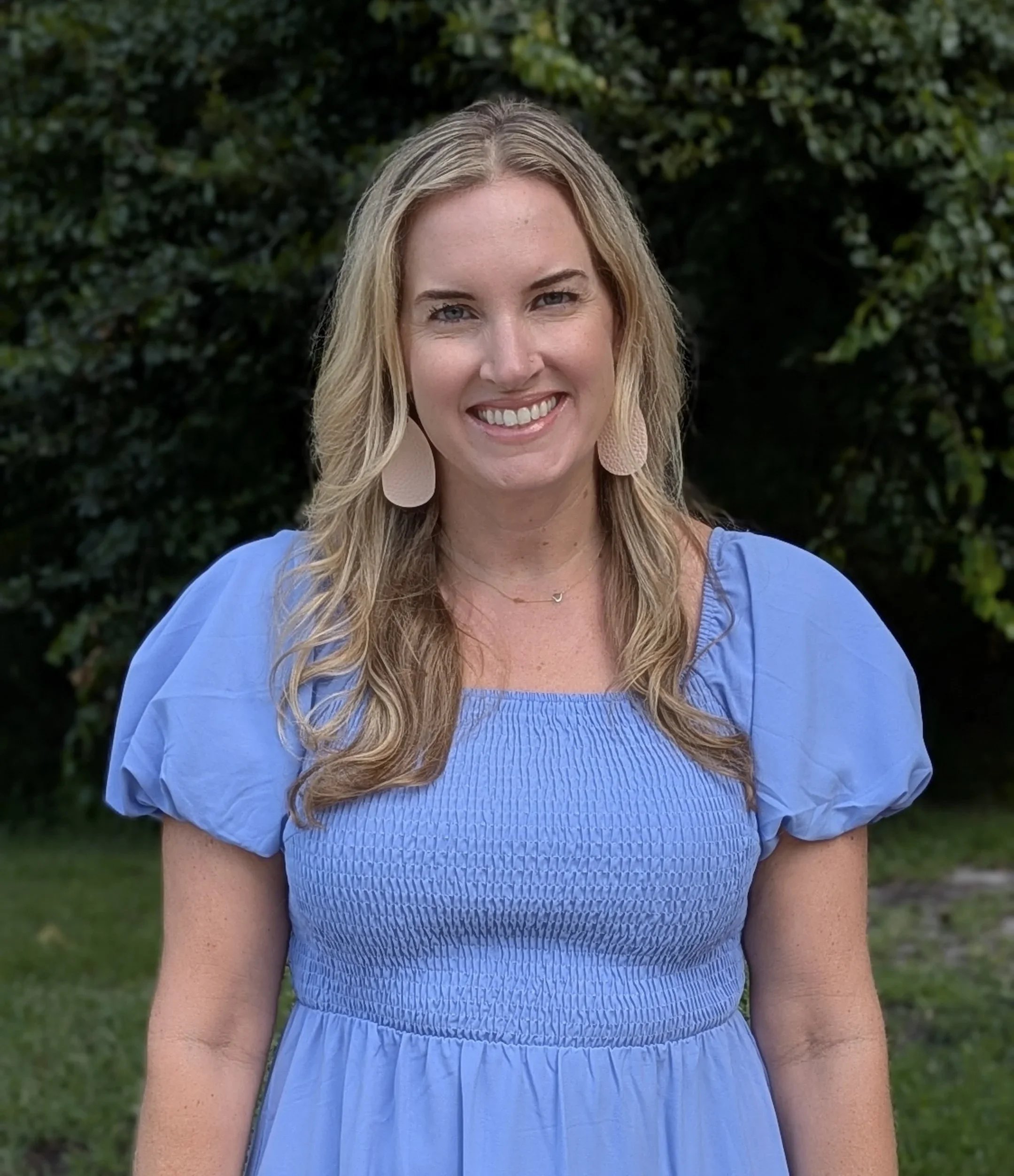 A woman with blonde, wavy hair and light-colored eyes wearing a light blue dress with puffed sleeves and large beige earrings, standing in front of green foliage and smiling.