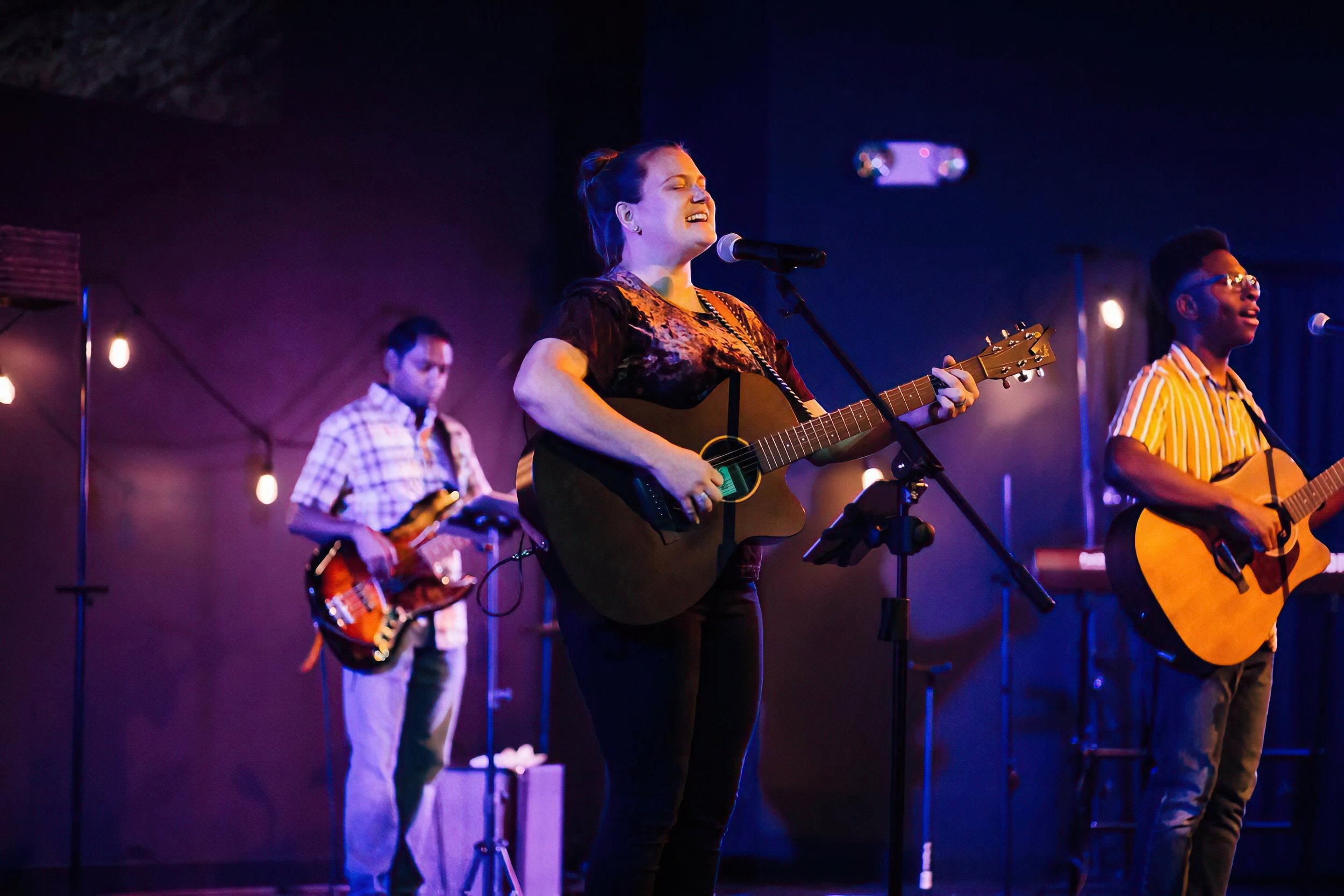 A woman singing and playing guitar on stage with two other musicians, one playing bass guitar, in a dimly lit venue with purple and blue lighting.