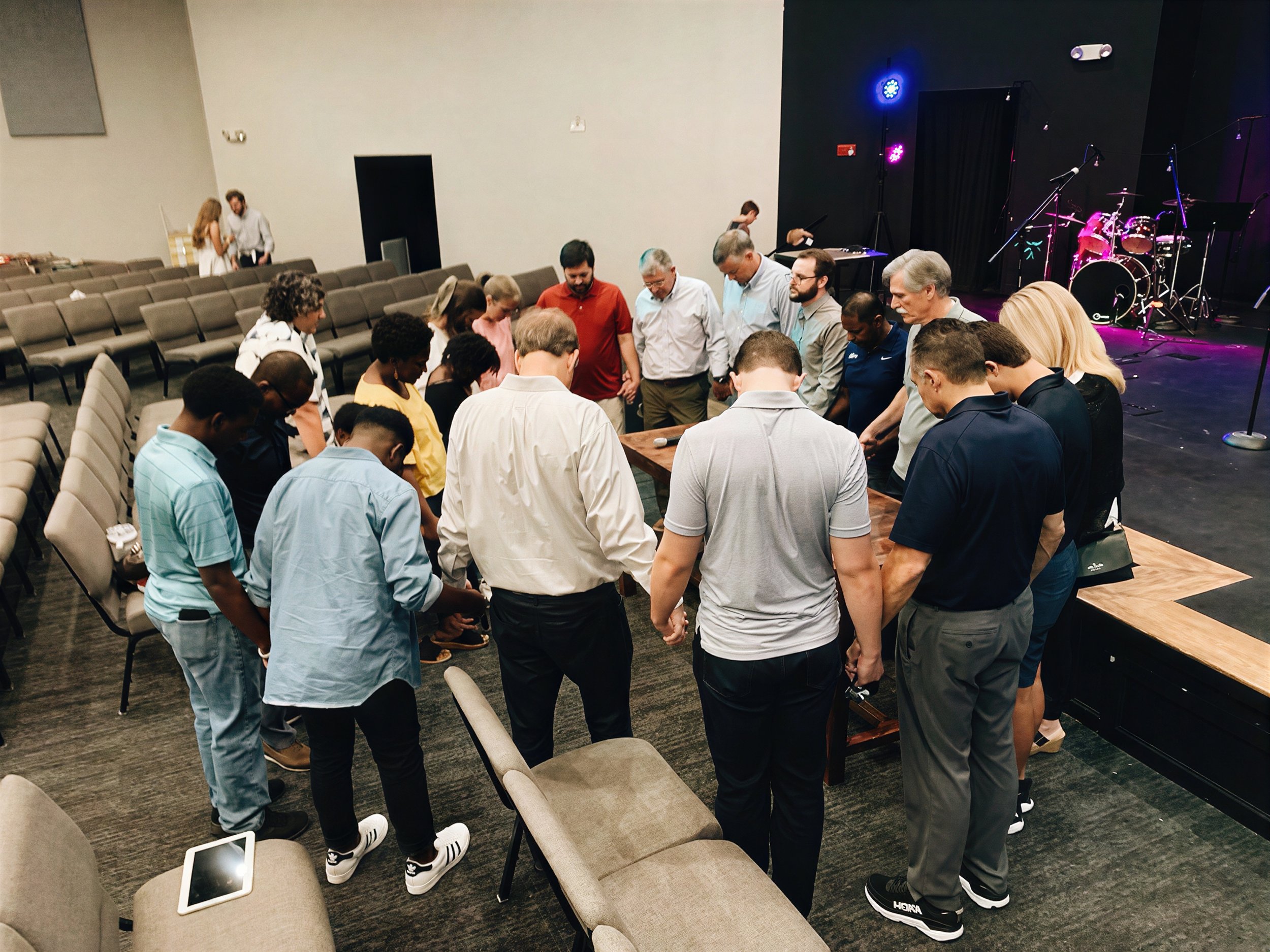 A group of diverse people standing in a circle with heads bowed in prayer or reflection in a room with empty chairs and a stage with musical instruments.