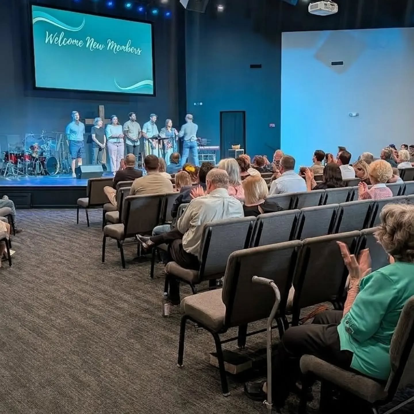 An indoor event with a group of people sitting in chairs, watching a stage with several individuals standing in front of a large screen that says "Welcome New Members". The stage has musical instruments to the side.