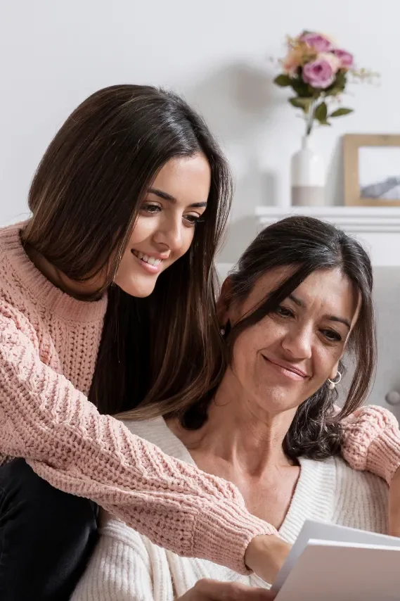 A young woman and an older woman sit together, looking at a tablet, smiling, in a cozy room with a vase of pink flowers on a shelf in the background.