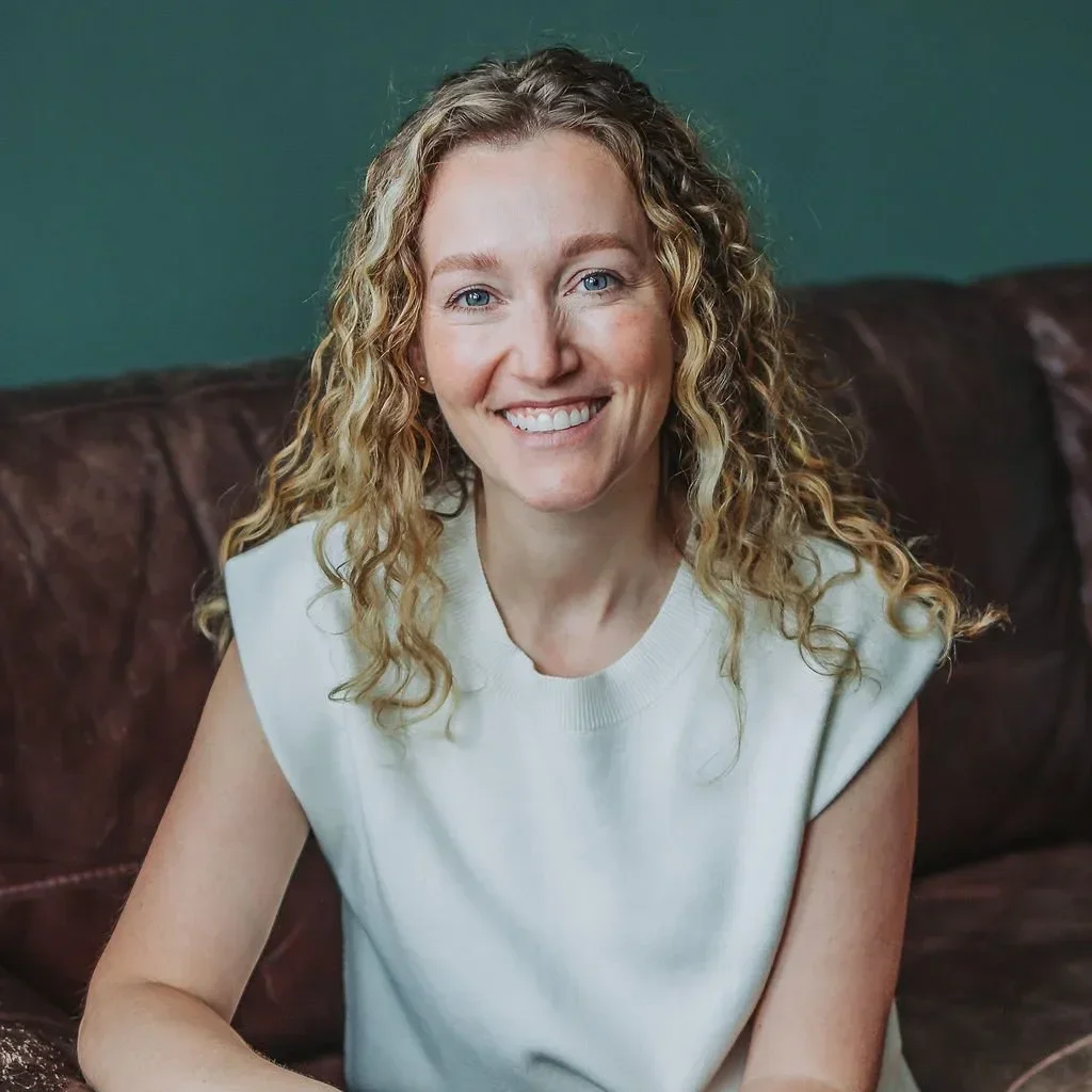 Dr. Angela Koenig with curly blonde hair wearing a sleeveless white top, smiling while sitting on a brown leather couch with a teal wall background.