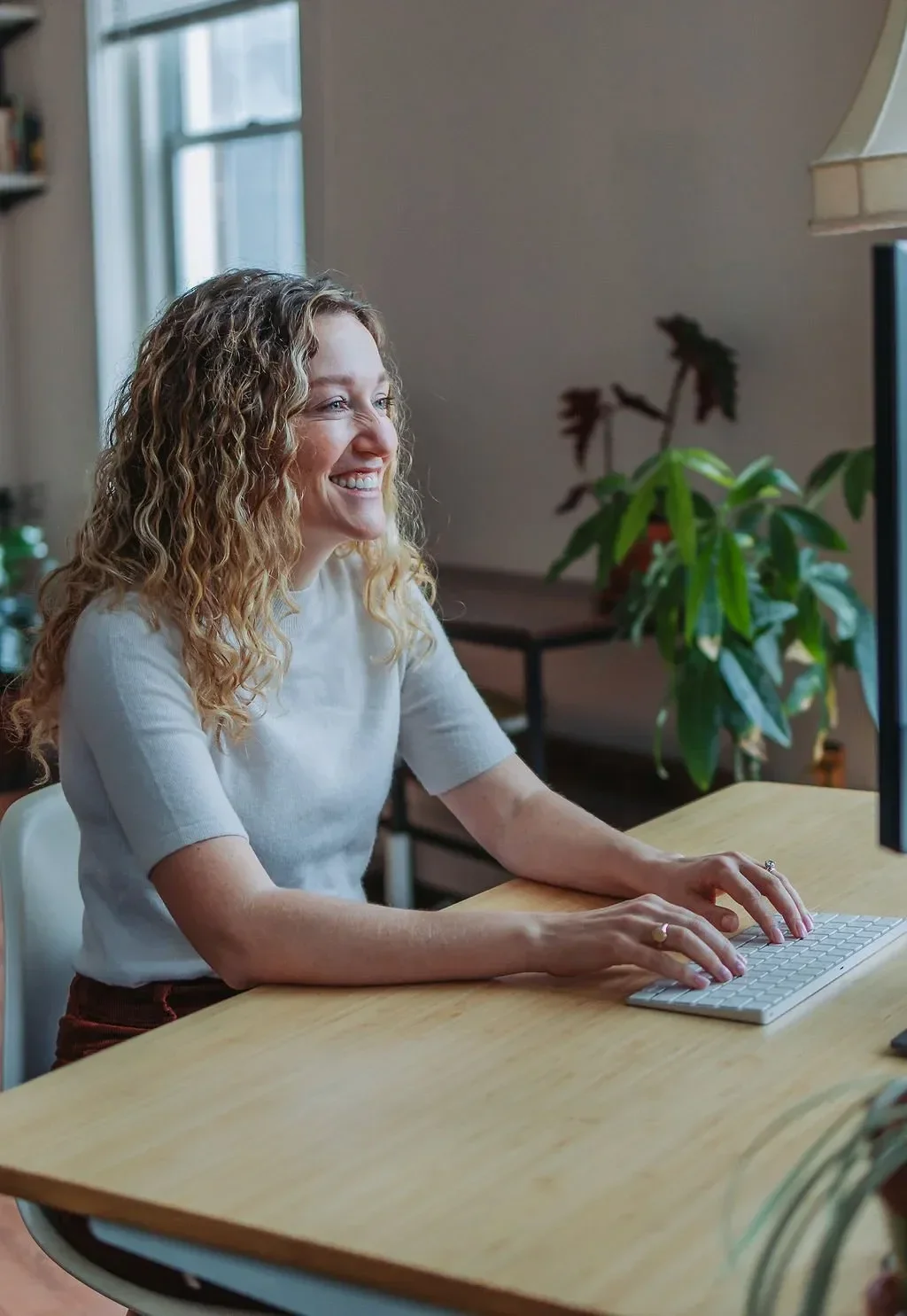 Dr. Angela Koenig with curly blonde hair working at a desk, smiling while using a computer keyboard in a bright, cozy room with a large window and houseplants.
