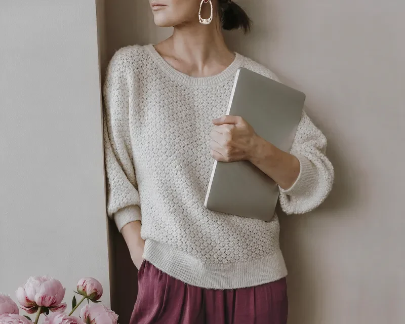 Woman wearing a sweater and earrings holding a silver laptop, standing by a wall near a window with pink peonies in front.