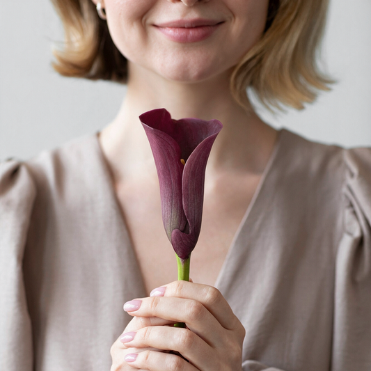 A woman holding a dark purple calla lily flower with both hands, smiling softly, wearing a beige blouse.