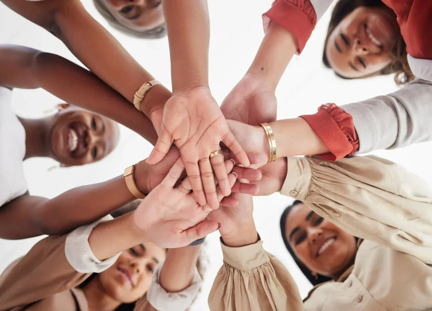 A diverse group of people is gathered in a circle, stacking their hands together in the center, smiling and showing unity and teamwork.