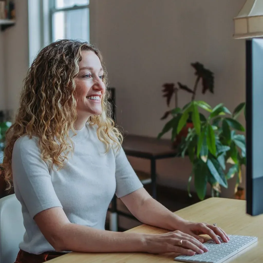 Dr. Angela Koenig with curly blonde hair working at a desk, smiling while looking at a computer screen, in a bright room with plants and a window.