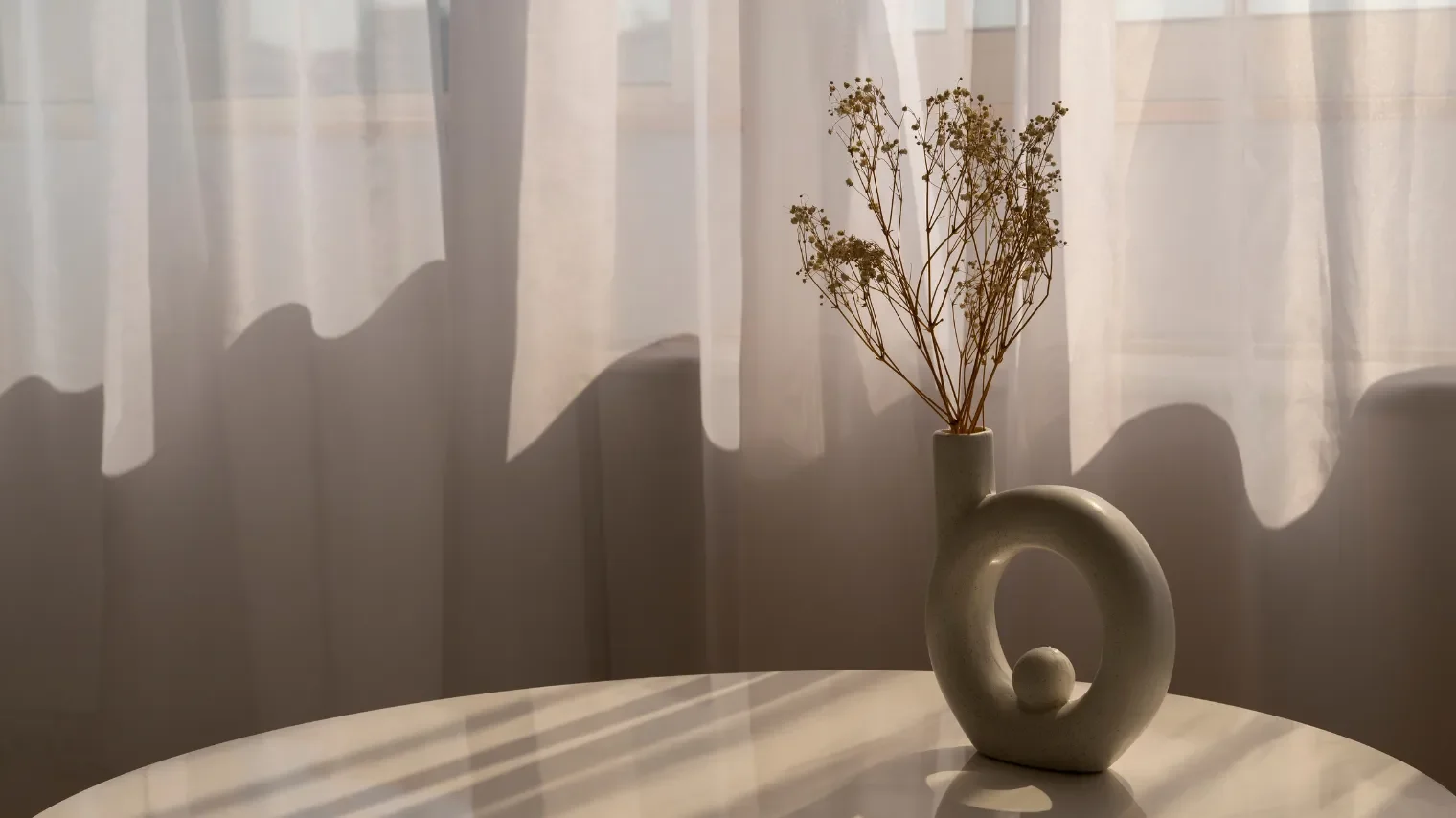 A decorative ceramic vase with a loop handle and a small sphere inside, holding dried flowers, placed on a white marble table near sheer curtains with sunlight creating shadows.
