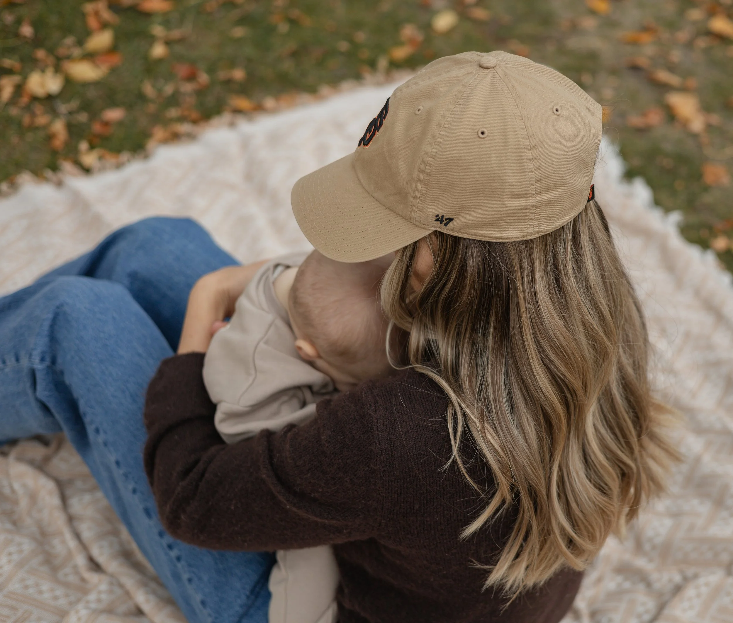 A person wearing a tan baseball cap with black and orange embroidery sitting outdoors on a blanket with a child resting against them, surrounded by fallen autumn leaves.