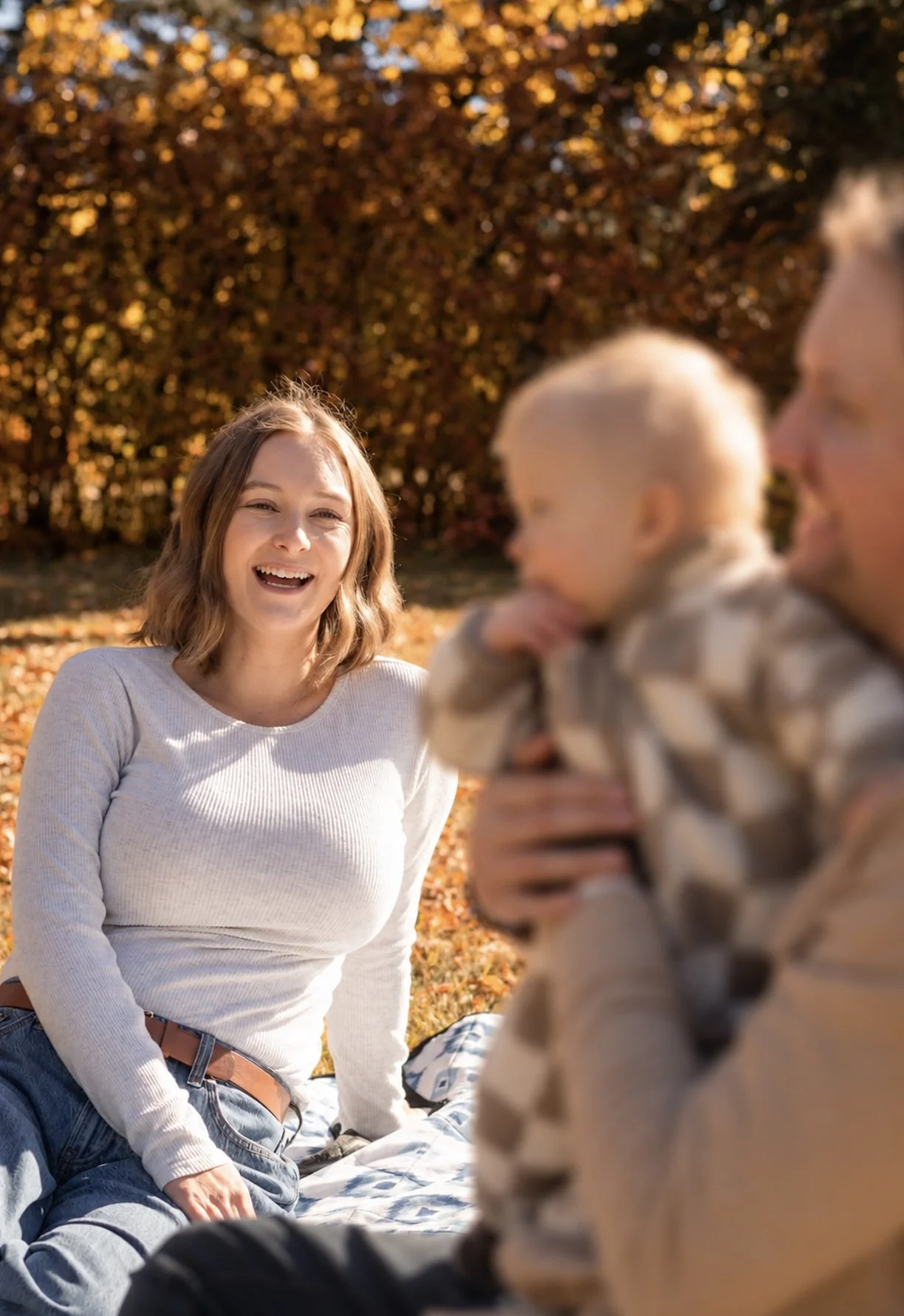A woman with shoulder-length brown hair wearing a white long-sleeve shirt, sitting on the ground outdoors, smiling at two young children. One child has blonde hair and is blur, and the other child, also blonde with shorter hair, is being held by an a