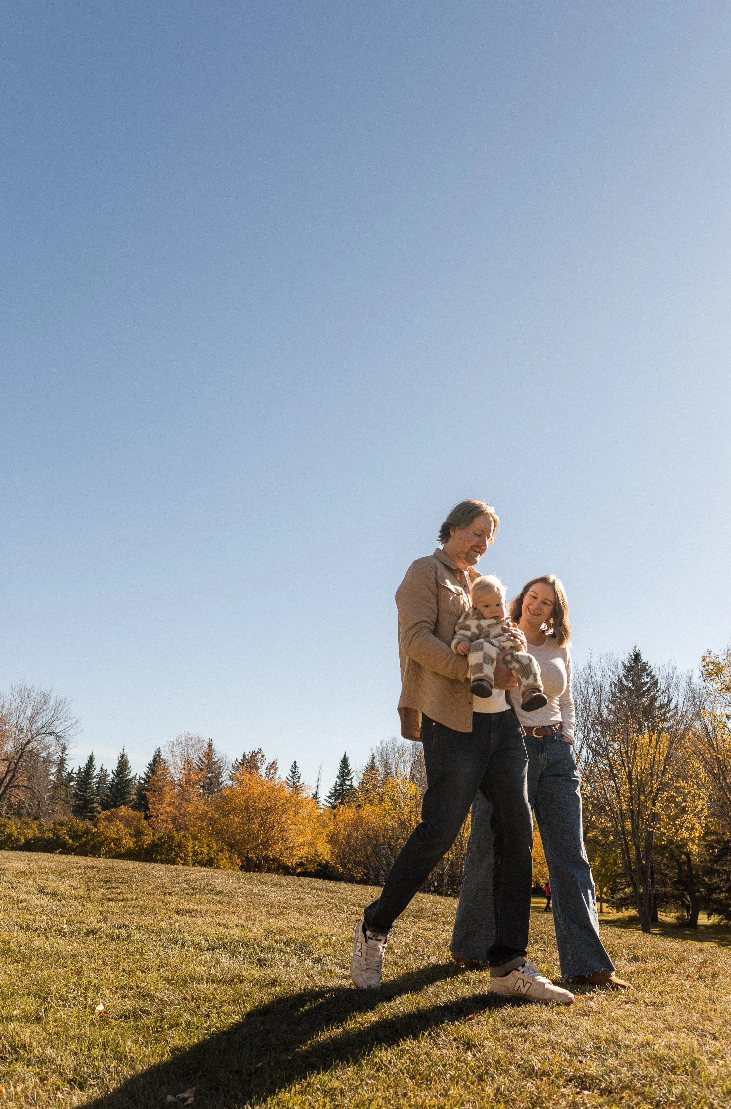 A family walking outside in a park with colorful autumn trees in the background on a sunny day. Location is Baker Park in Calgary, Alberta