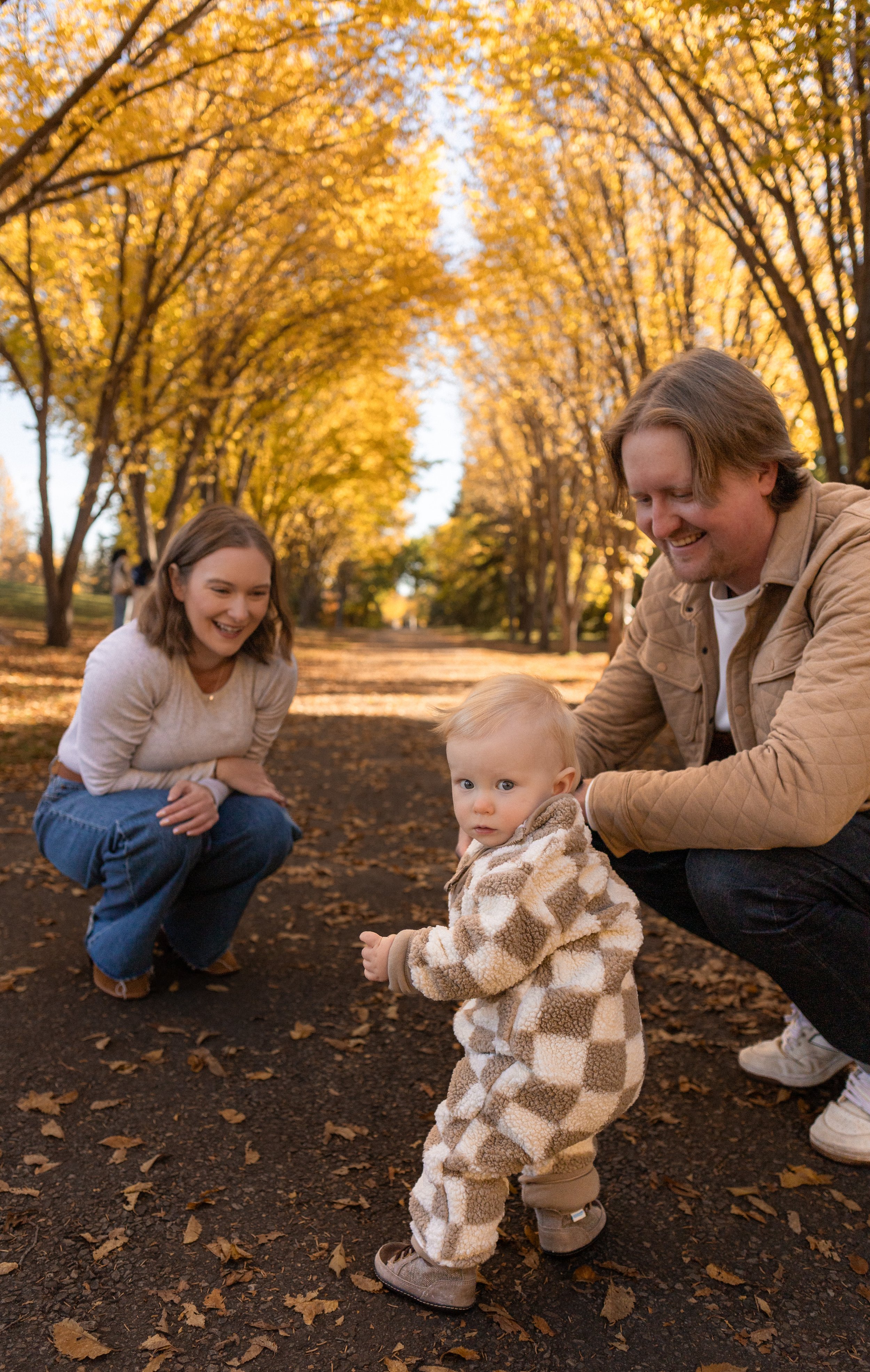 A family enjoying a walk in a park during autumn, with golden leaves on the trees and fallen on the ground. A woman, man, and young child are visible, with the child in the foreground wearing a cozy, patterned outfit.