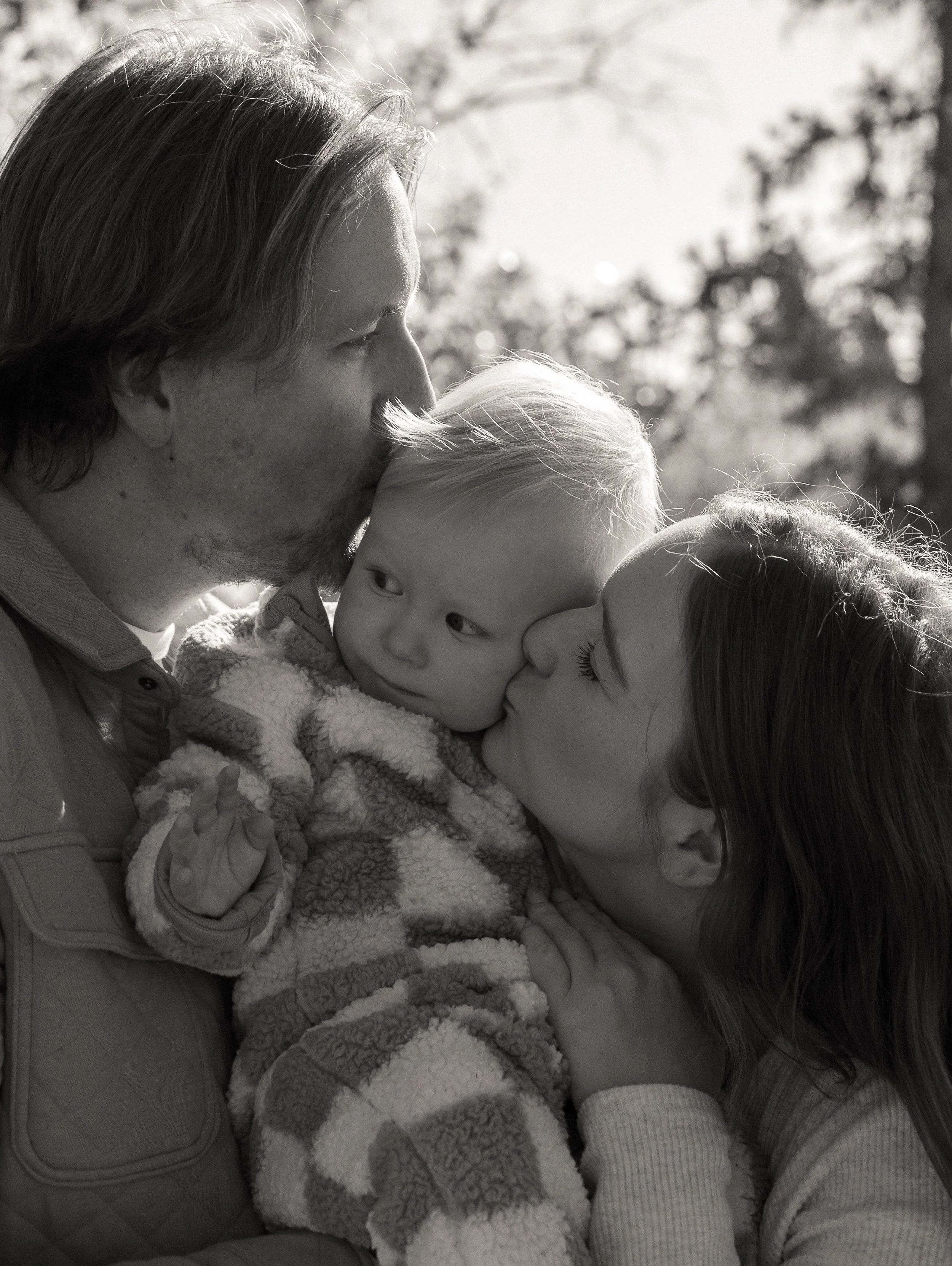 A black and white photo of a family of three, including a man, woman, and child, sharing a tender moment outdoors, with the man giving the child a kiss on the forehead and the woman embracing the child.