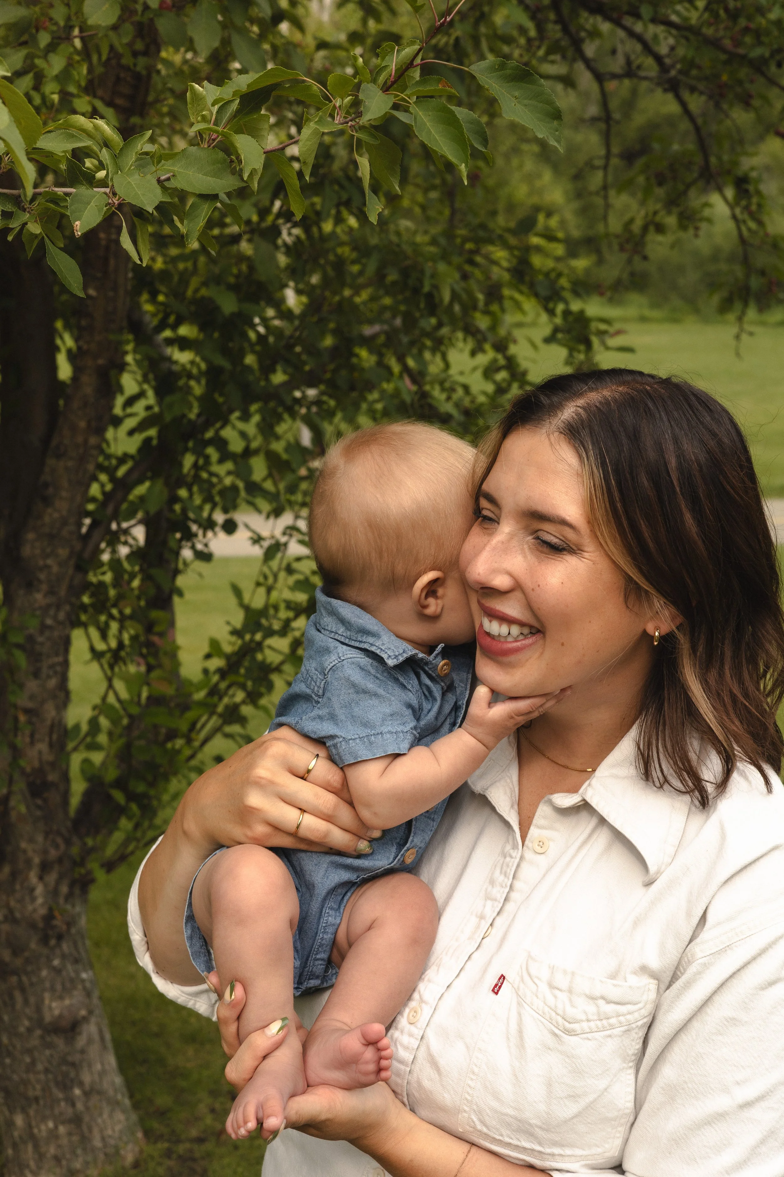 A woman with shoulder-length brown hair smiling and holding a young child outdoors near a tree with green leaves, in a grassy area.  Location is Confederation Park in Calgary, Alberta