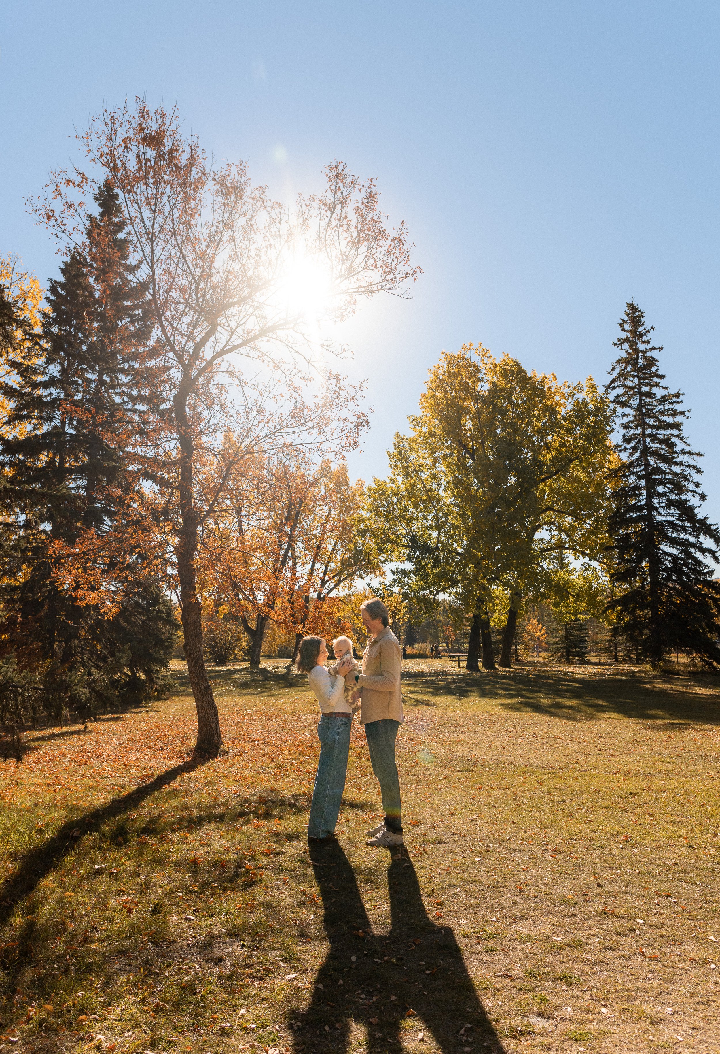 A family of three stands in a park with fall foliage, sunlight shining through trees, and long shadows on the ground. Location is Baker Park in Calgary, Alberta