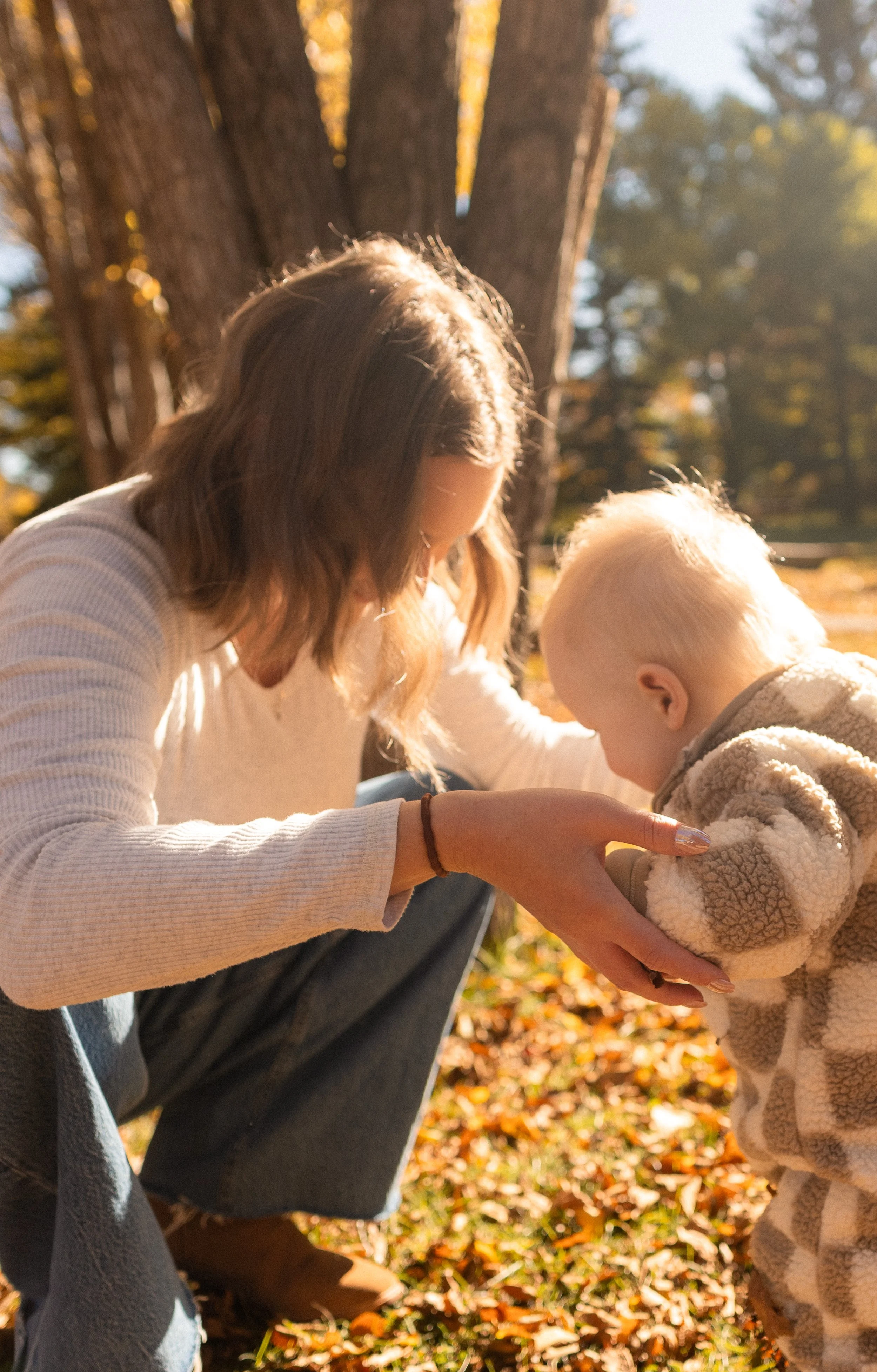 A woman with wavy brown hair and light skin crouches down outdoors in autumn, holding a young child with blonde hair and light skin who is wearing a beige fleece jacket, as they both look at something in her hands. They are near a large tree with ora