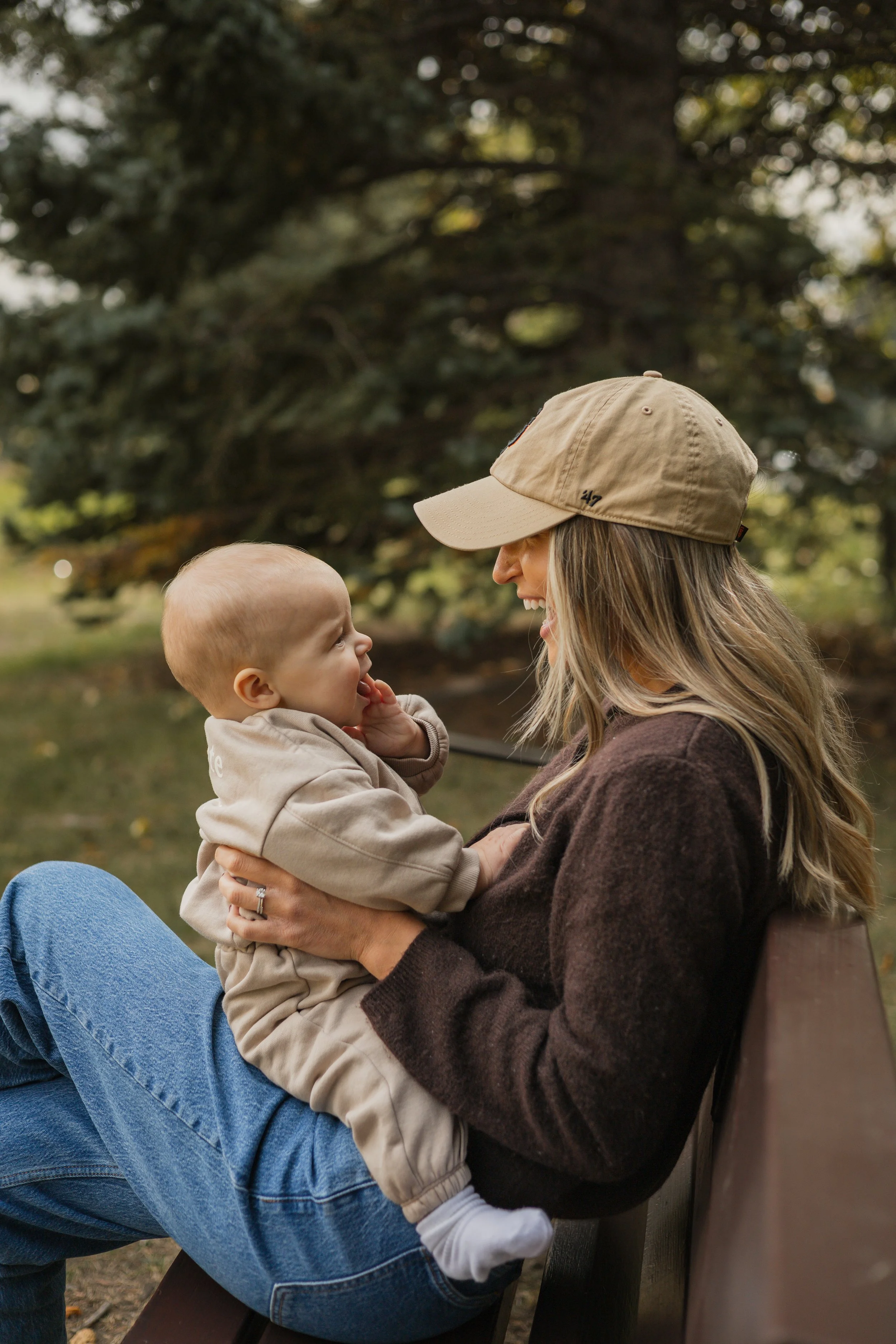 A woman sitting on a park bench holding a young child in her lap. They are smiling at each other, with trees in the background. The woman is wearing a tan cap and a brown sweater, while the child is in a beige jacket.