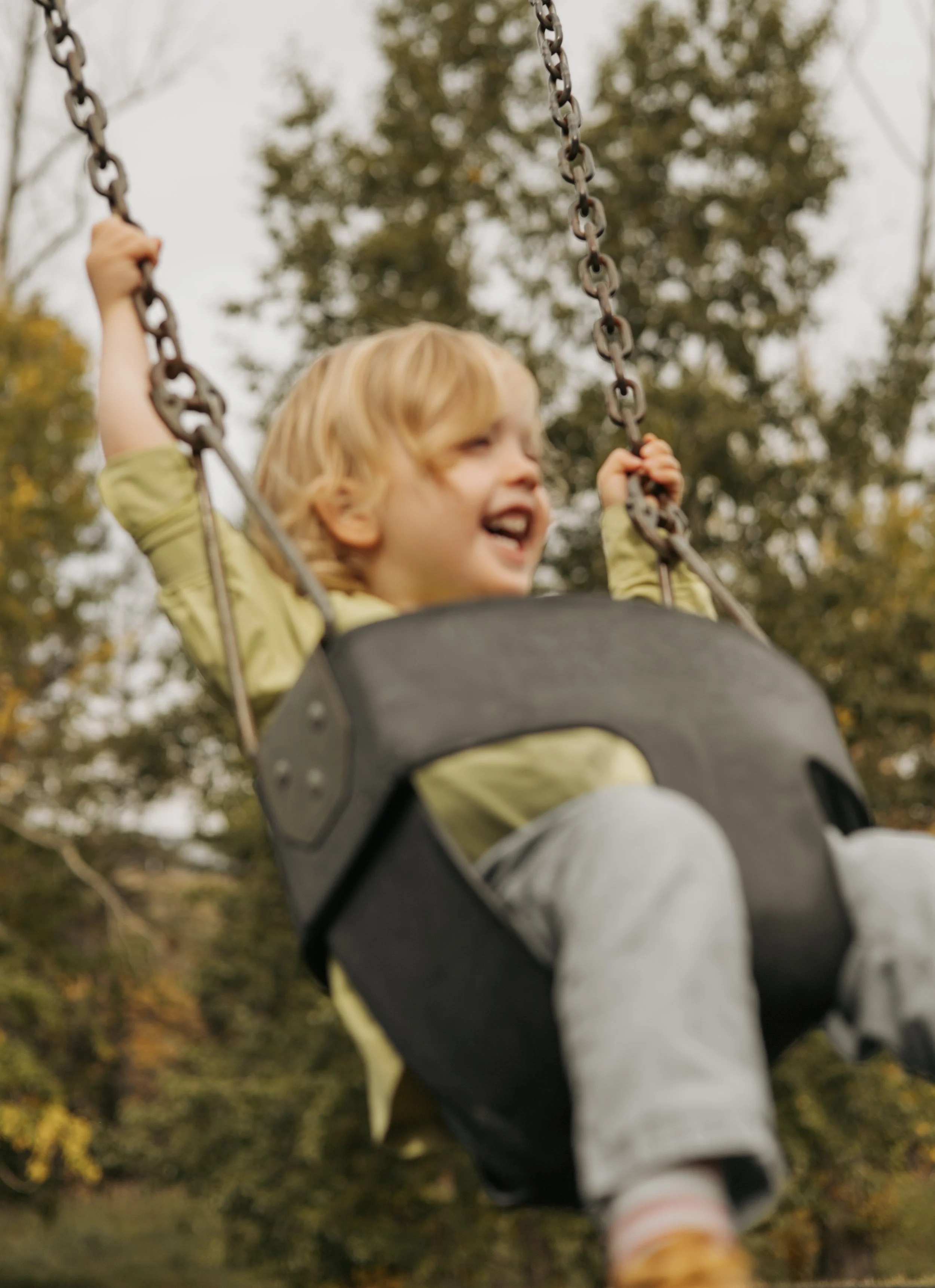 A young child with blonde hair smiling in a baby swing outdoors with trees in the background.