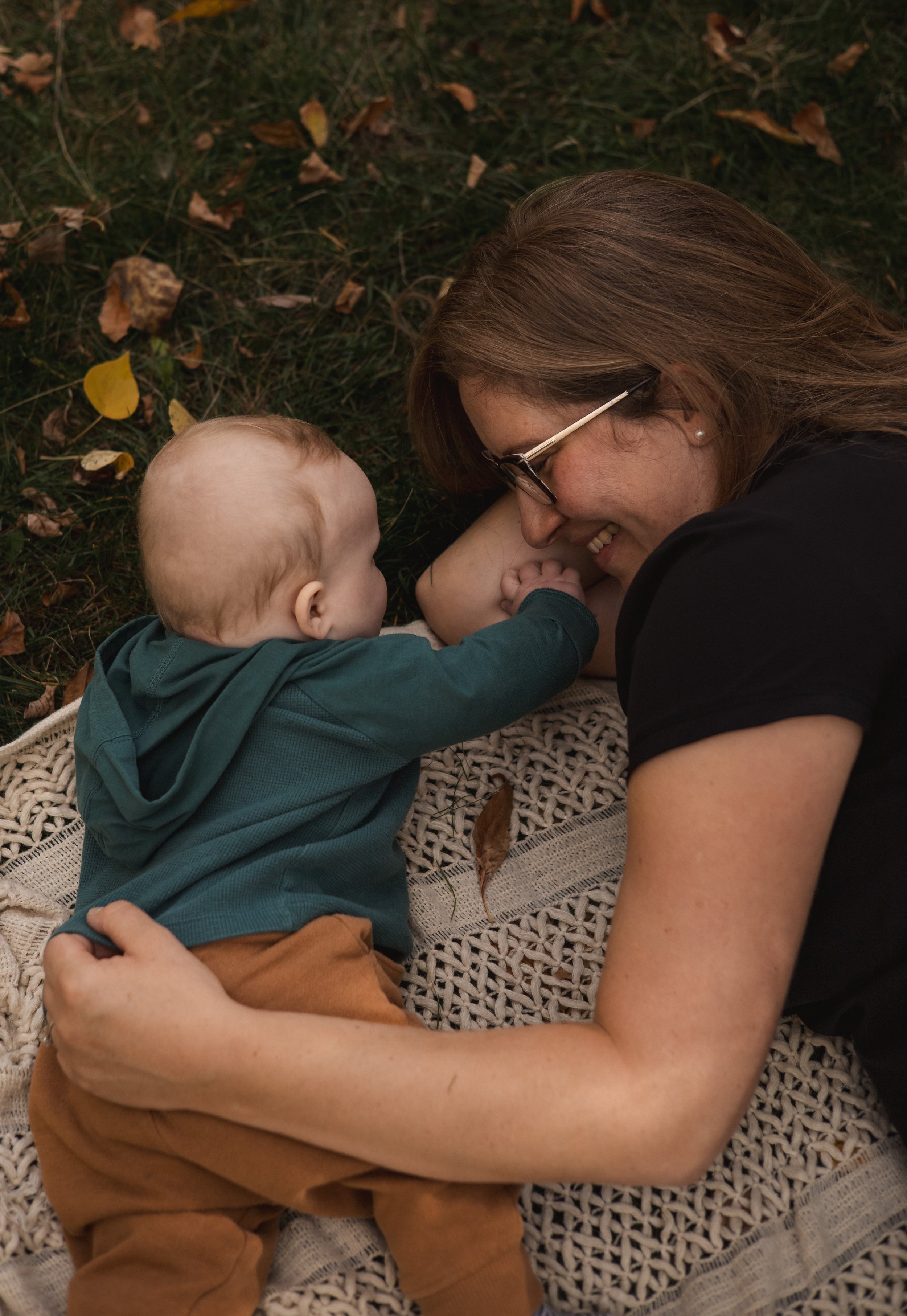 A woman with long brown hair and glasses smiling at a young child with blonde hair, lying on a blanket outdoors among fallen leaves.