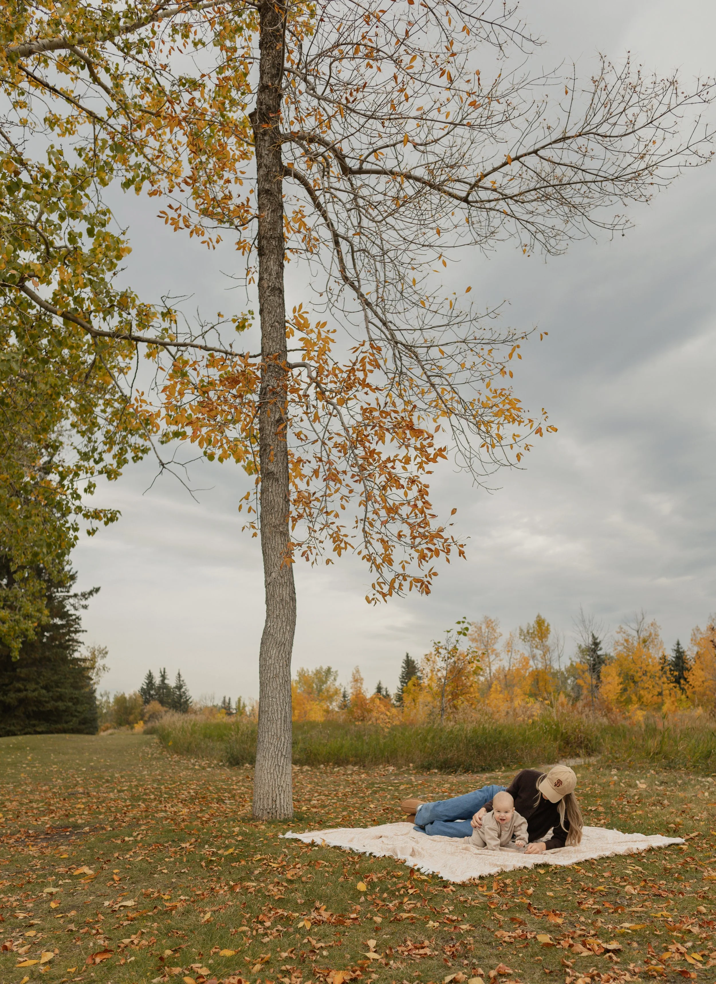 A woman lying on a blanket in a park with autumn-colored trees in the background, playing with a smiling baby amidst fallen leaves.