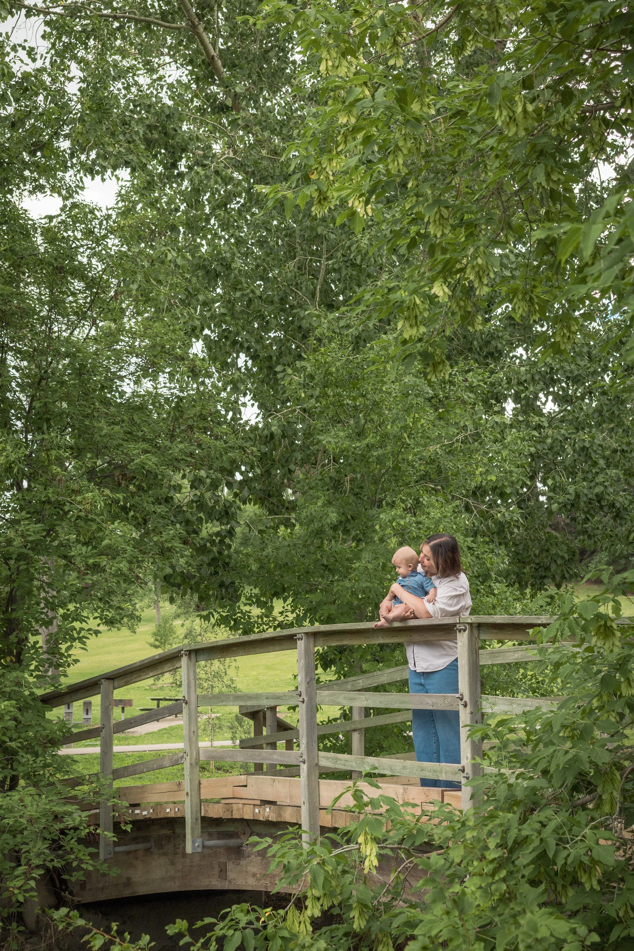 A woman holding a baby on a wooden bridge in a park with green trees and grass.  Location is Confederation Park in Calgary, Alberta