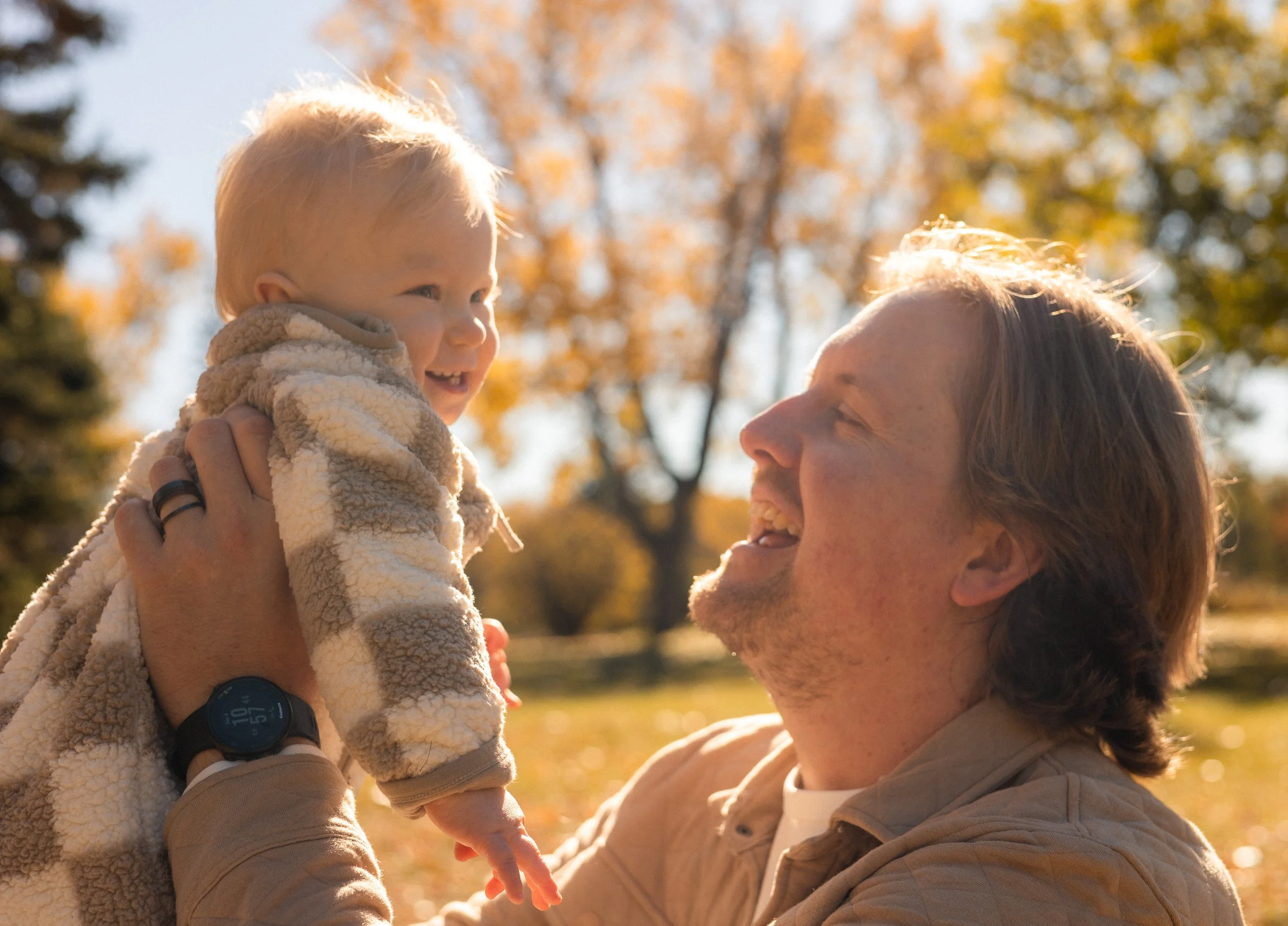 A woman is holding up a toddler in a park with autumn trees in the background. Both are smiling and enjoying the sunny day.