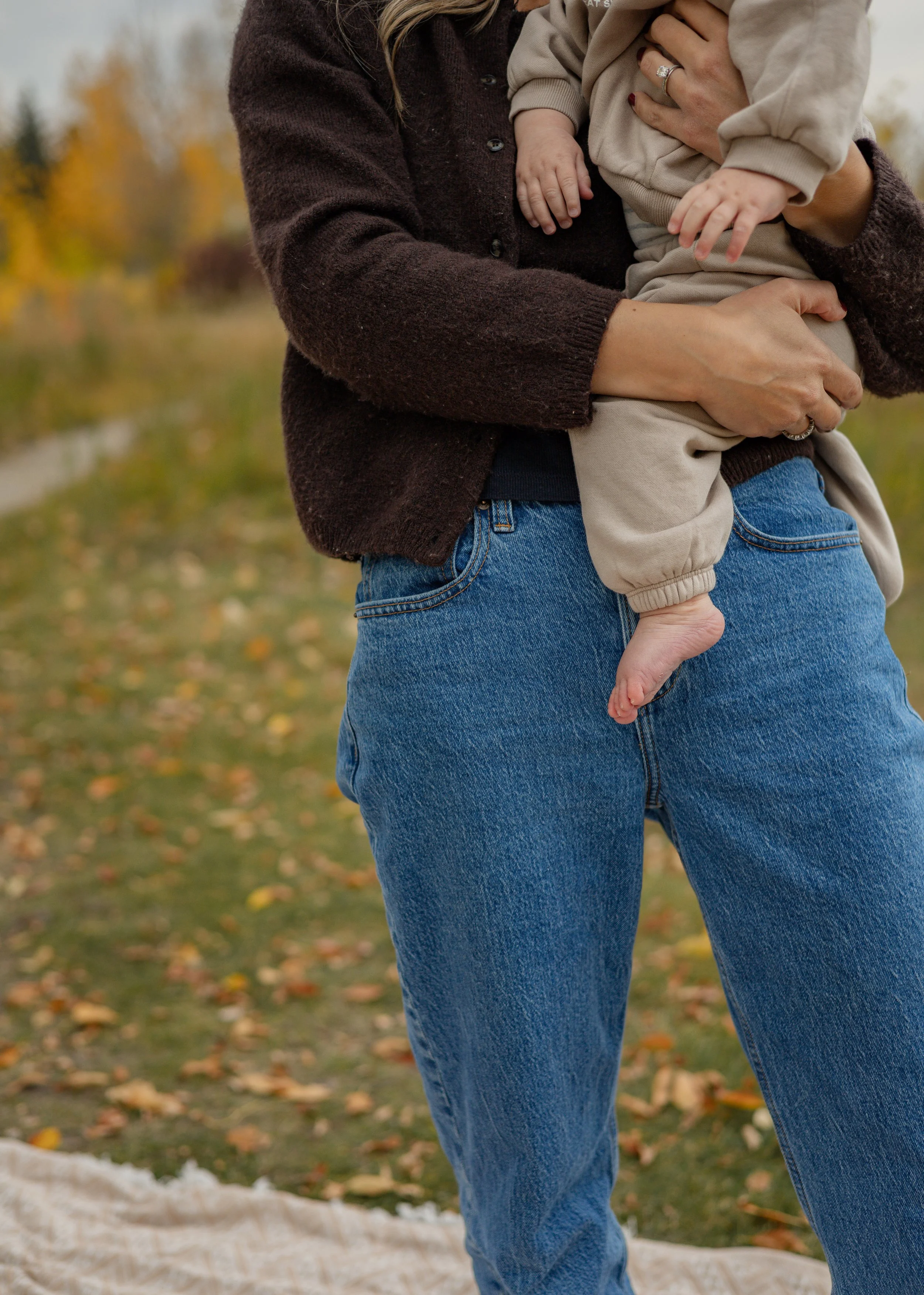 A woman holding a small child outdoors in a park with fall foliage in the background.