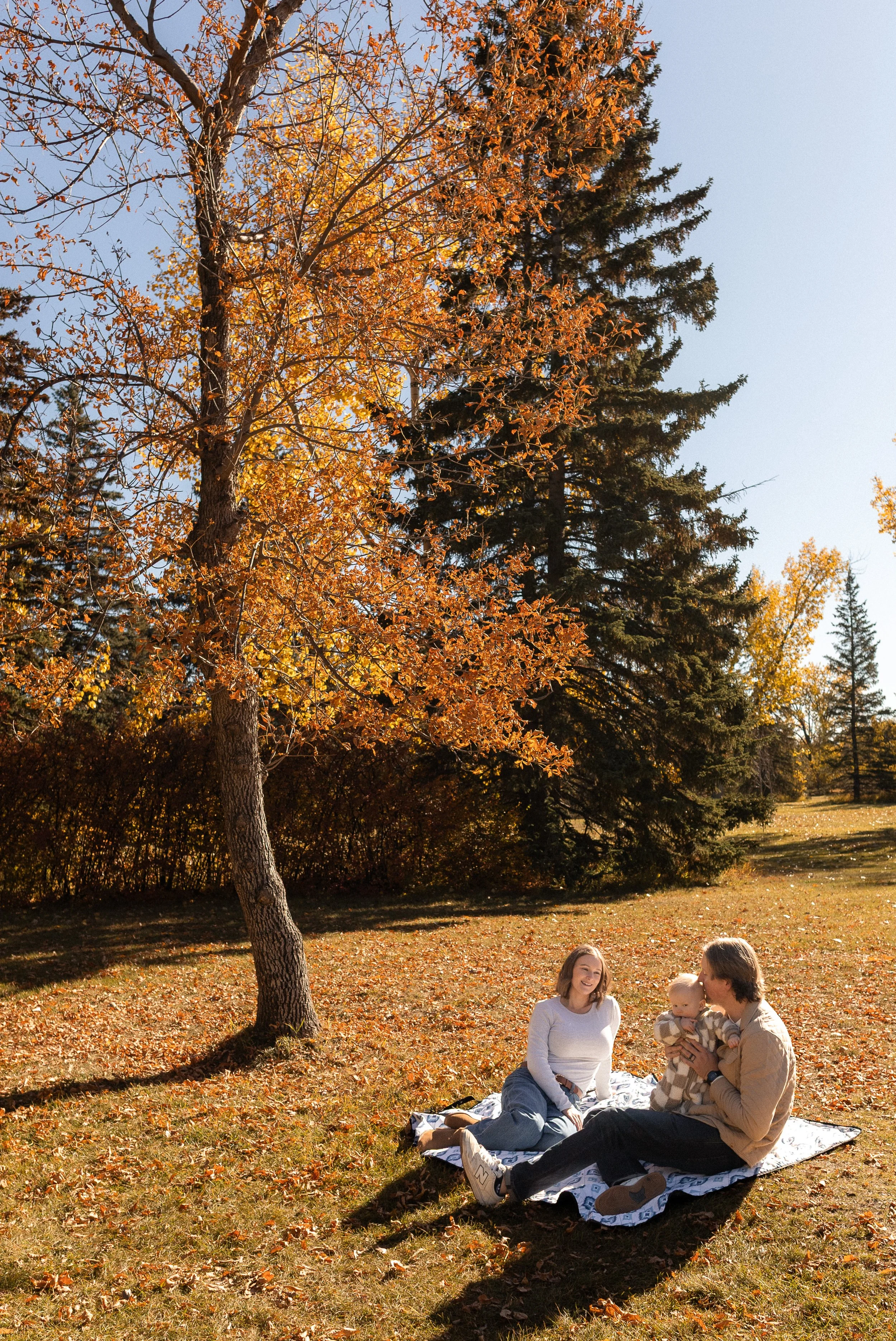 A family sitting on a blanket in a park during fall. A woman is smiling, a man is holding a baby, and colorful autumn leaves cover the ground and trees. The sky is clear and blue.