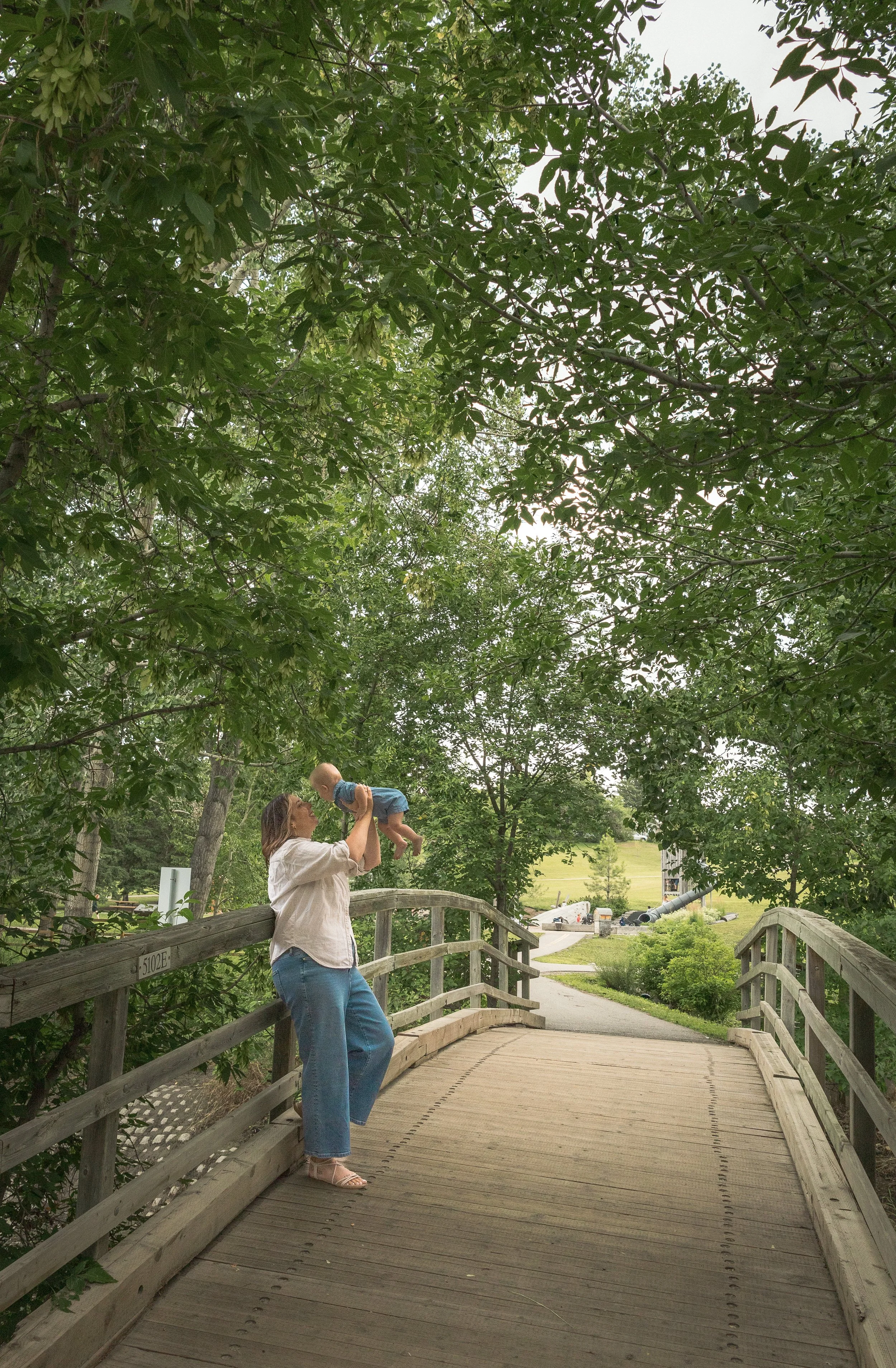 A woman in blue jeans and pink sandals is holding a child up in the air on a wooden bridge in a park with trees and open grassy fields in the background.