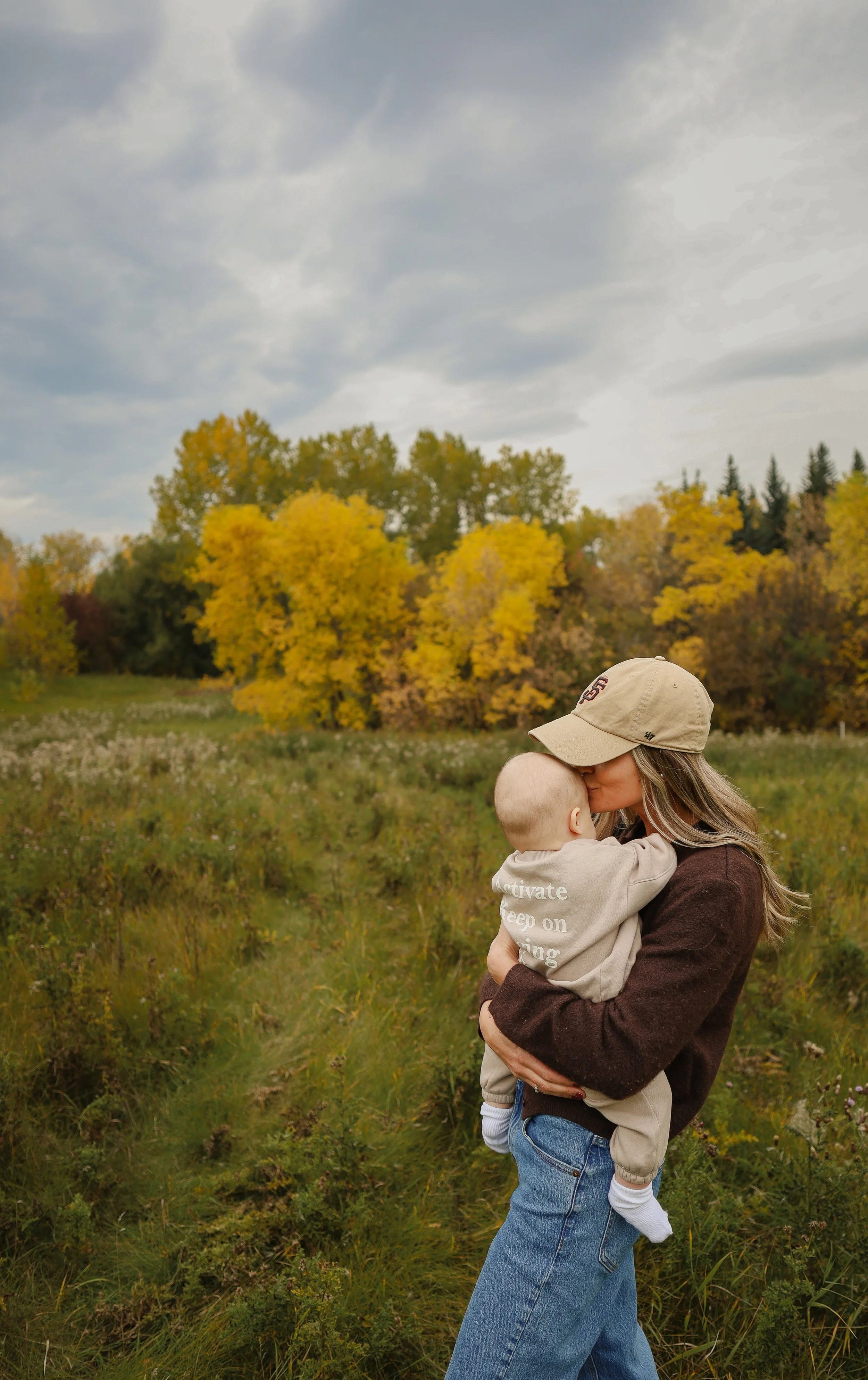 A woman with long blonde hair, wearing a beige cap and brown sweater, is holding a young child in a field of grass and wildflowers. The child is dressed in a beige sweatshirt and jeans, and they are sharing an affectionate moment outdoors during autu