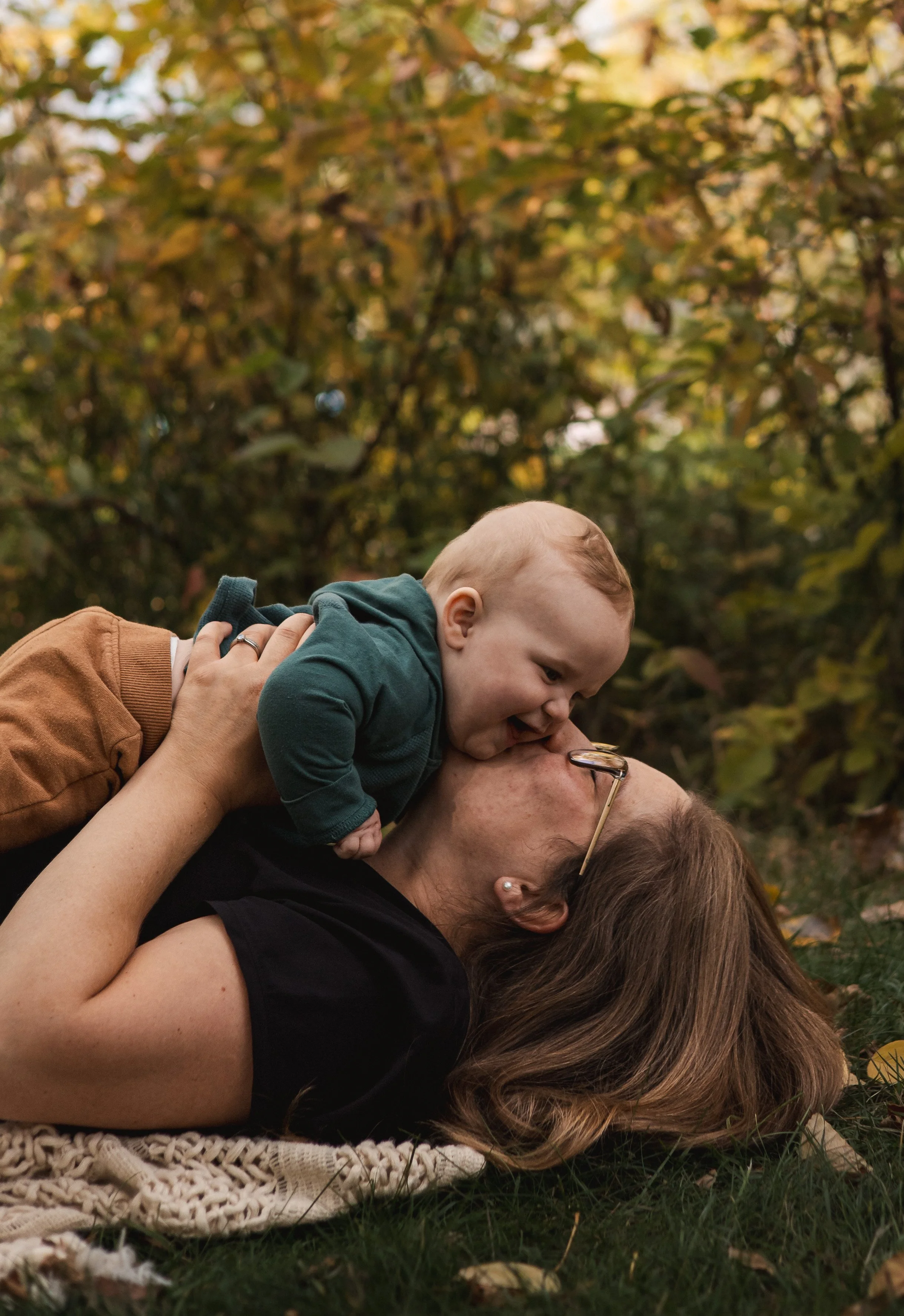 A family of three on a blanket lying on the grass, outdoors during autumn, with fall foliage in the background. The child is on top of the woman, both are smiling. Location is Bowness Park in Calgary, Alberta