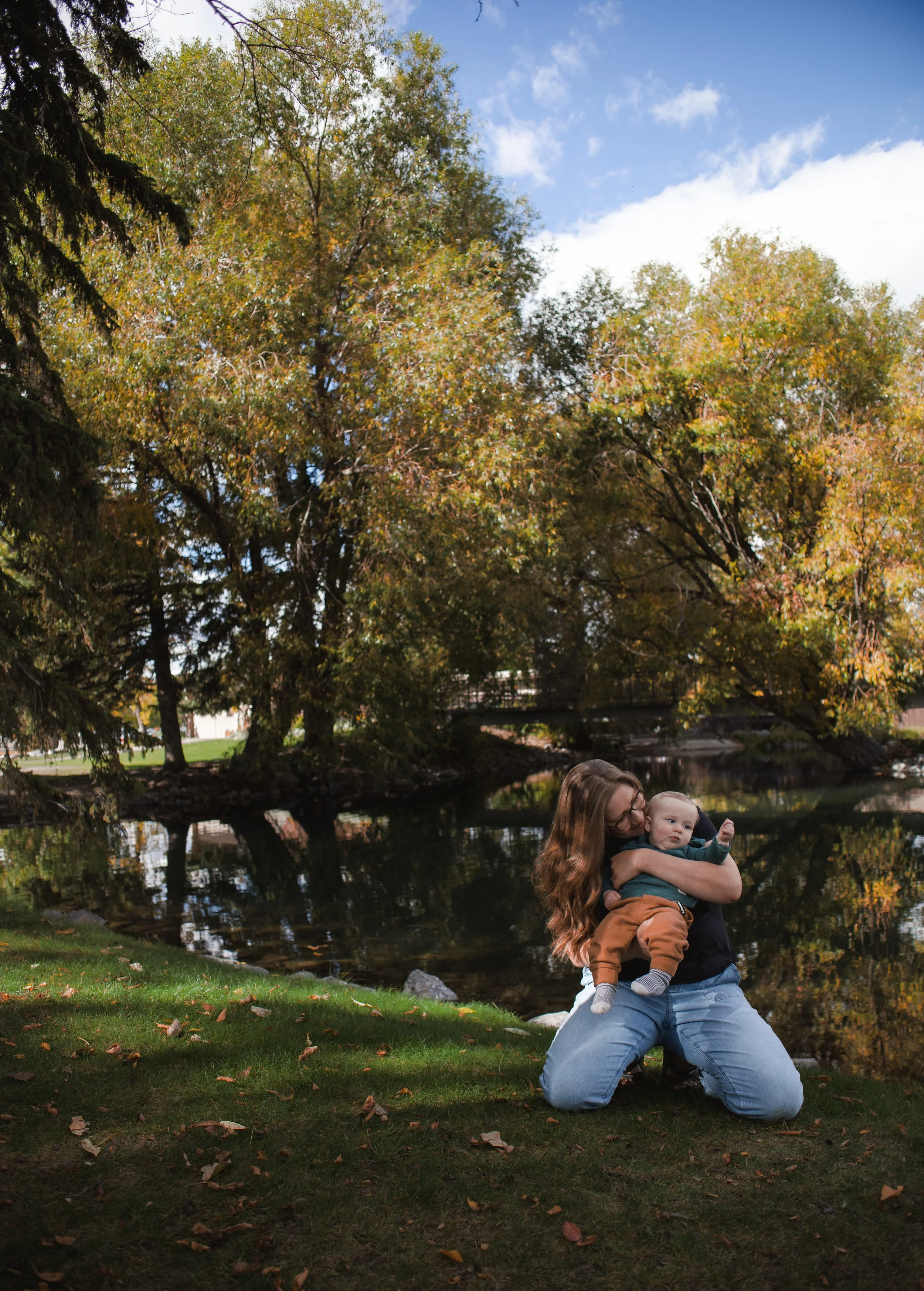 A woman with long hair and glasses kneeling by a small pond, holding a young child in her arms. The woman is wearing a dark shirt and jeans, and the child is dressed in an orange and green outfit. The background features tall trees with autumn leaves